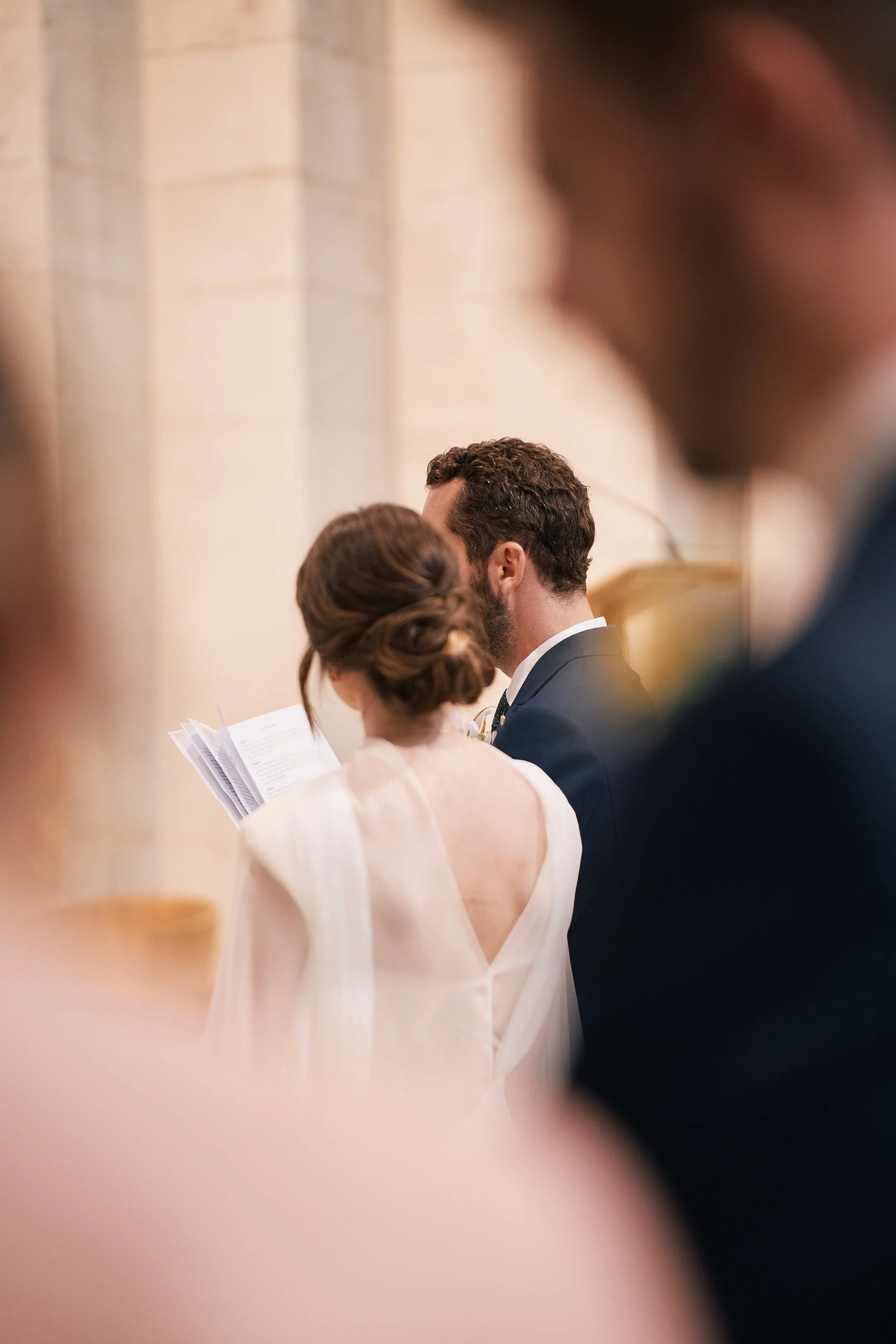 A bride and groom stand close during a wedding ceremony, surrounded by blurred guests.