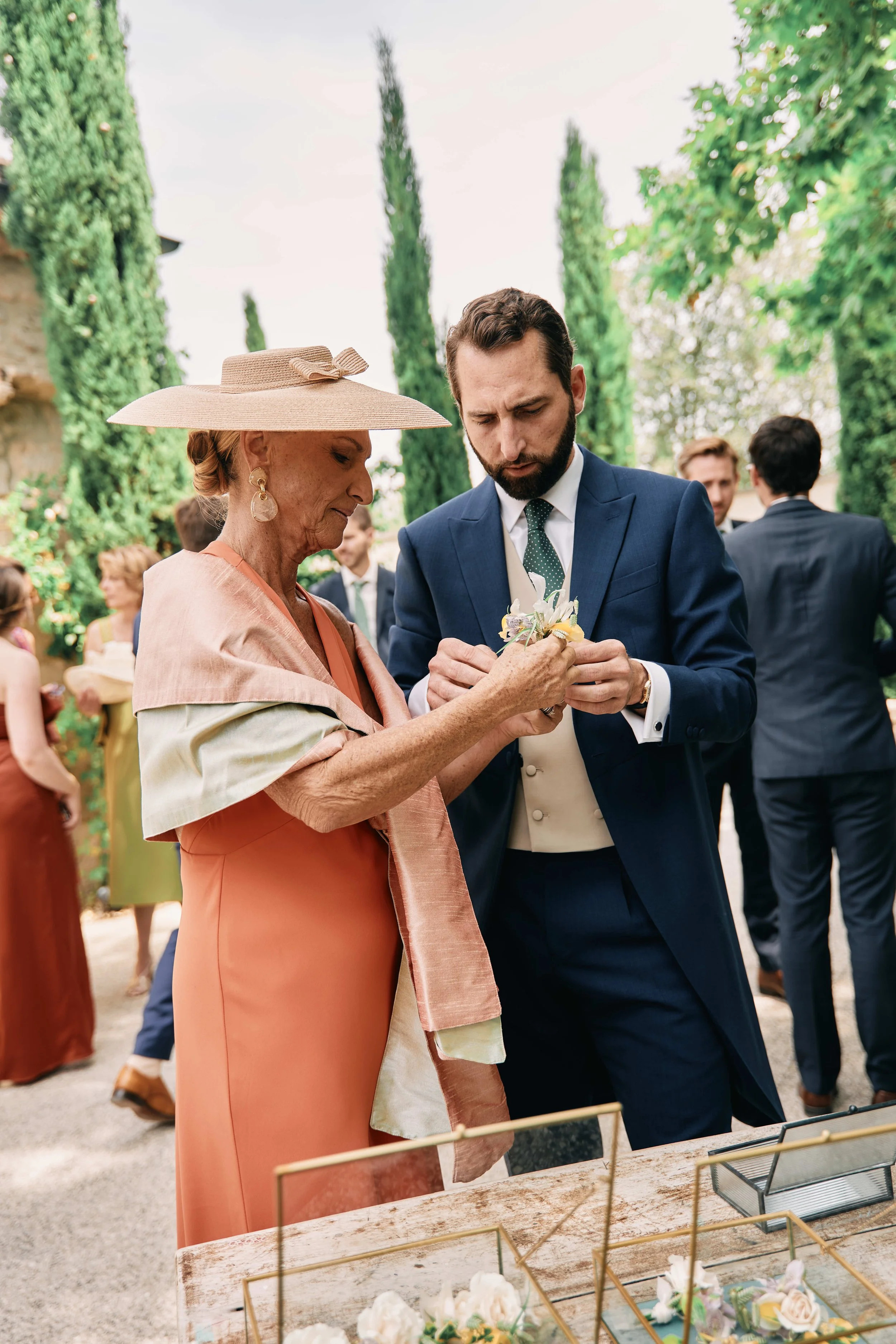 A young man, wearing a blue suit and green tie, helping an elderly woman in a peach dress and large wide-brimmed hat at an outdoor event with greenery in the background.