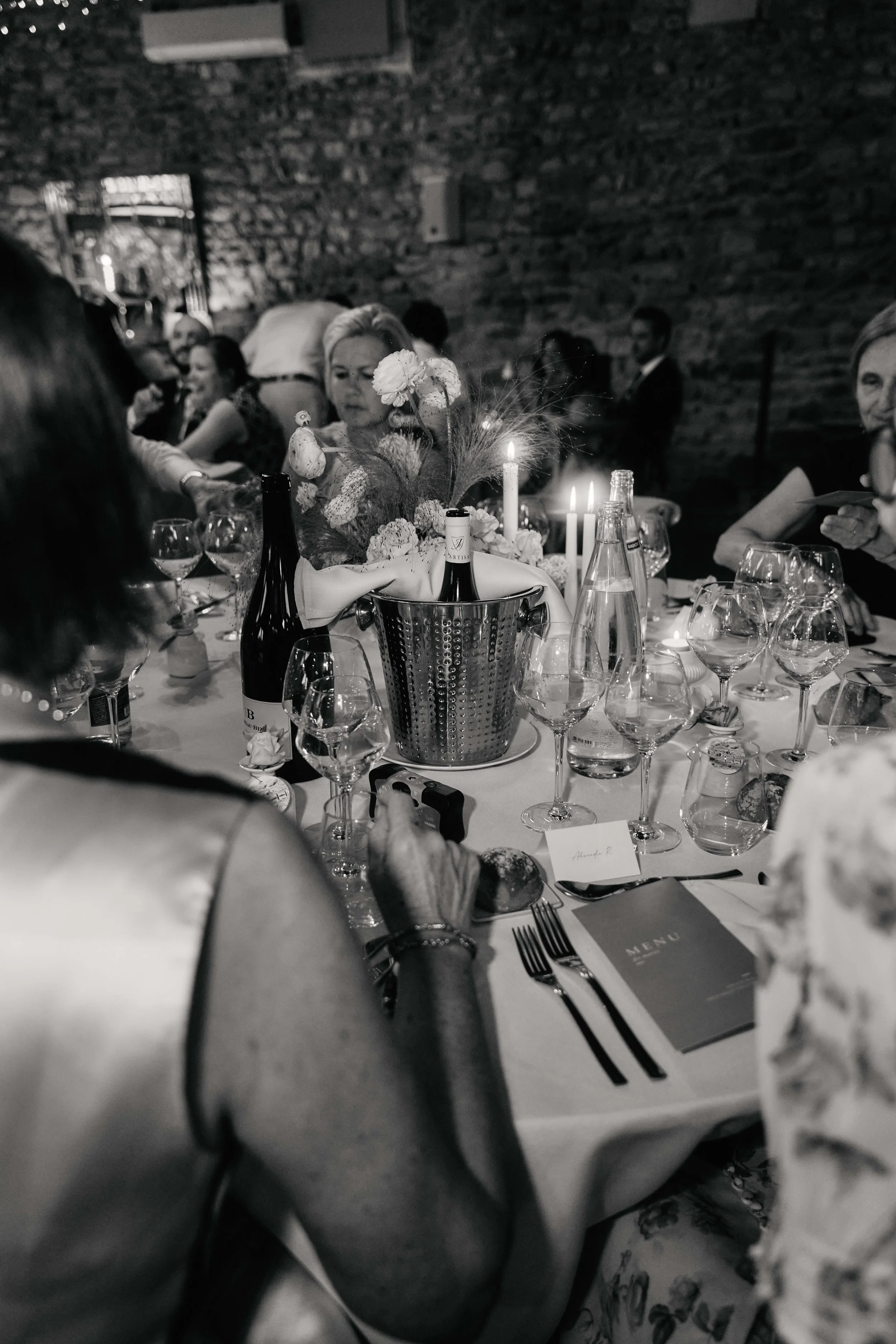 A black and white photo of a dining table at a formal event with multiple wine glasses, bottles, candles, and a menu card on a white tablecloth. There are several guests seated, engaged in conversation, with a brick wall in the background.