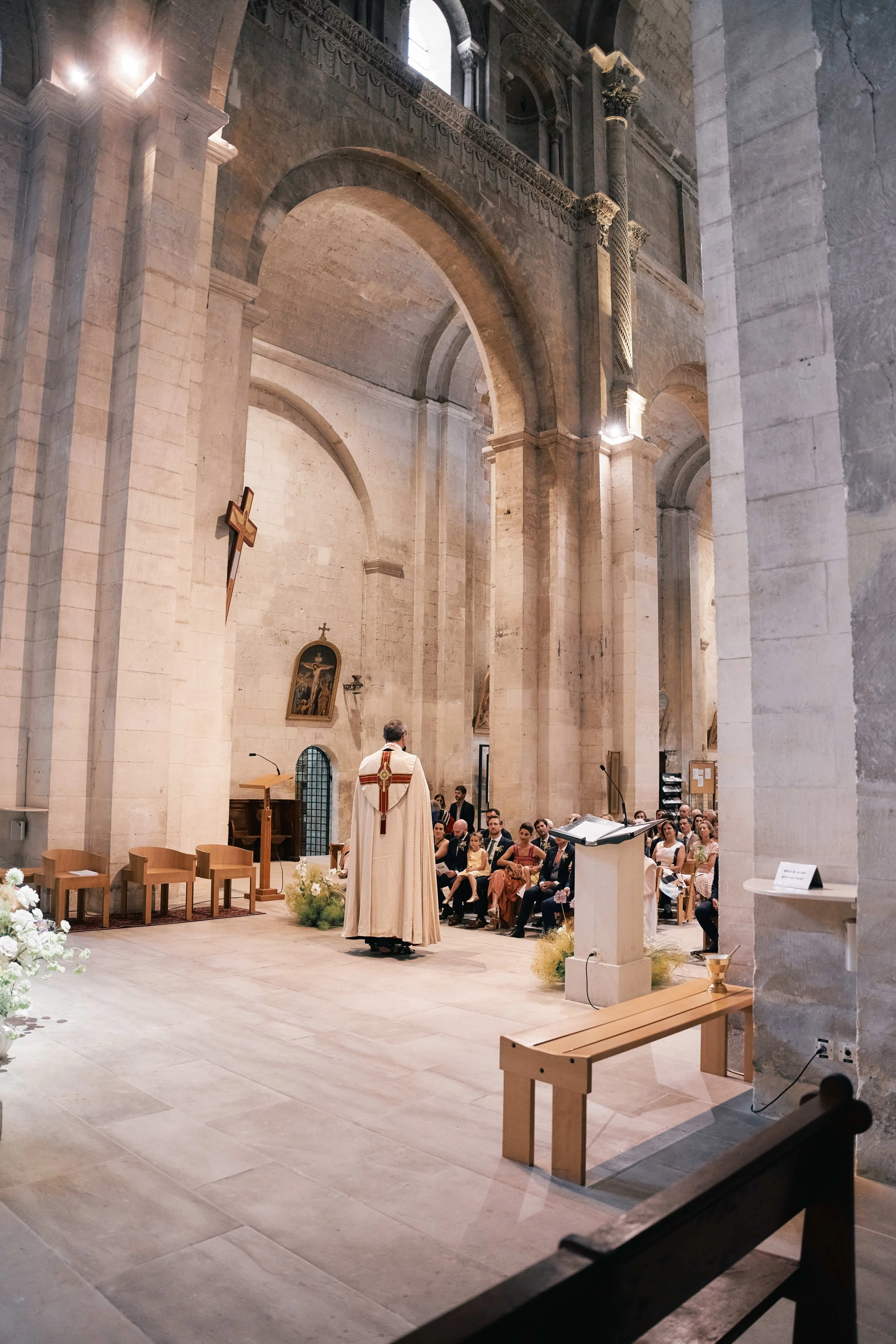 Inside a church during a wedding ceremony, with a priest officiating, guests seated, and religious symbols visible on the wall.