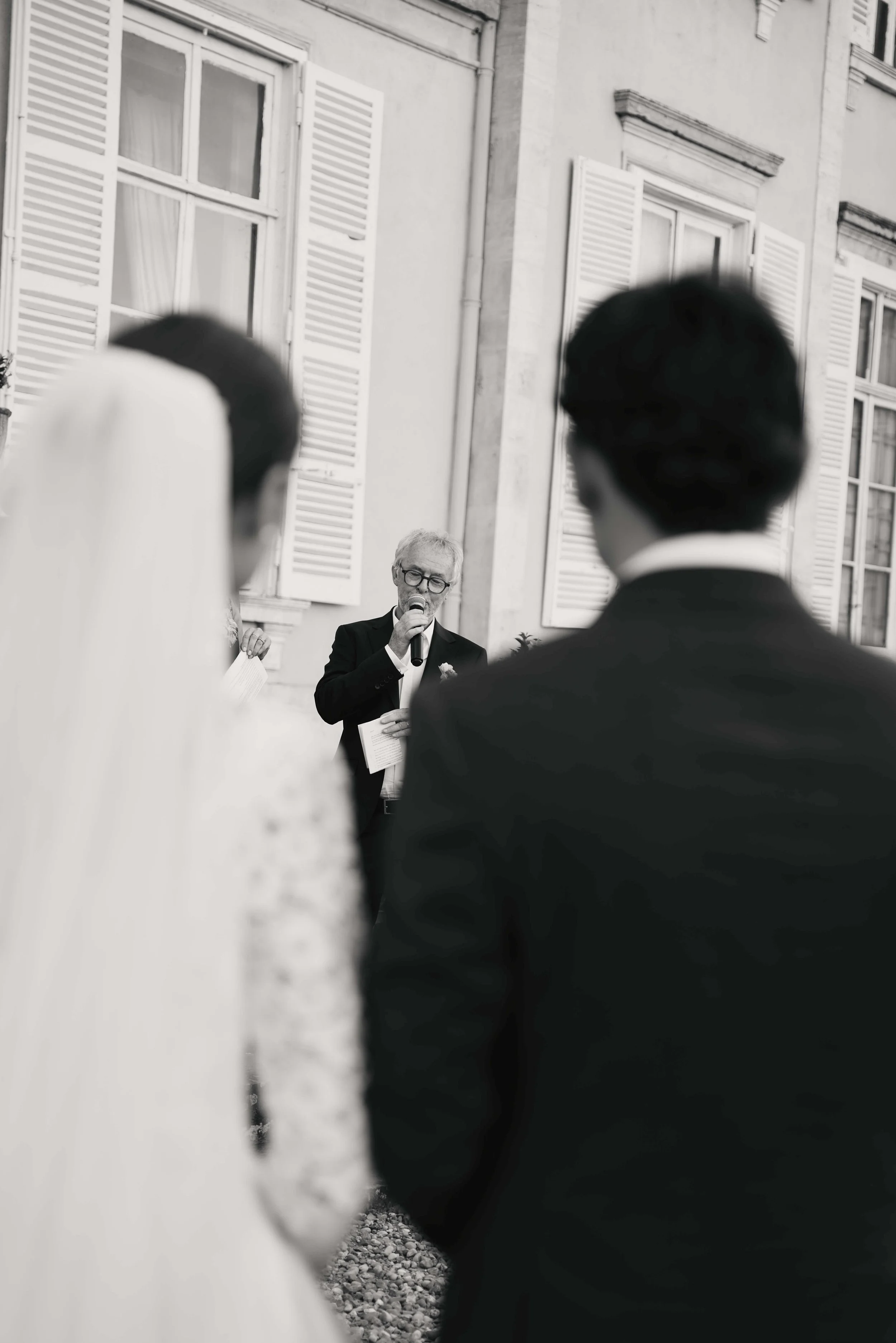Black and white photo of a man with glasses and a suit speaking into a microphone, standing in front of a building with windows and shutters, with two other people in the foreground.