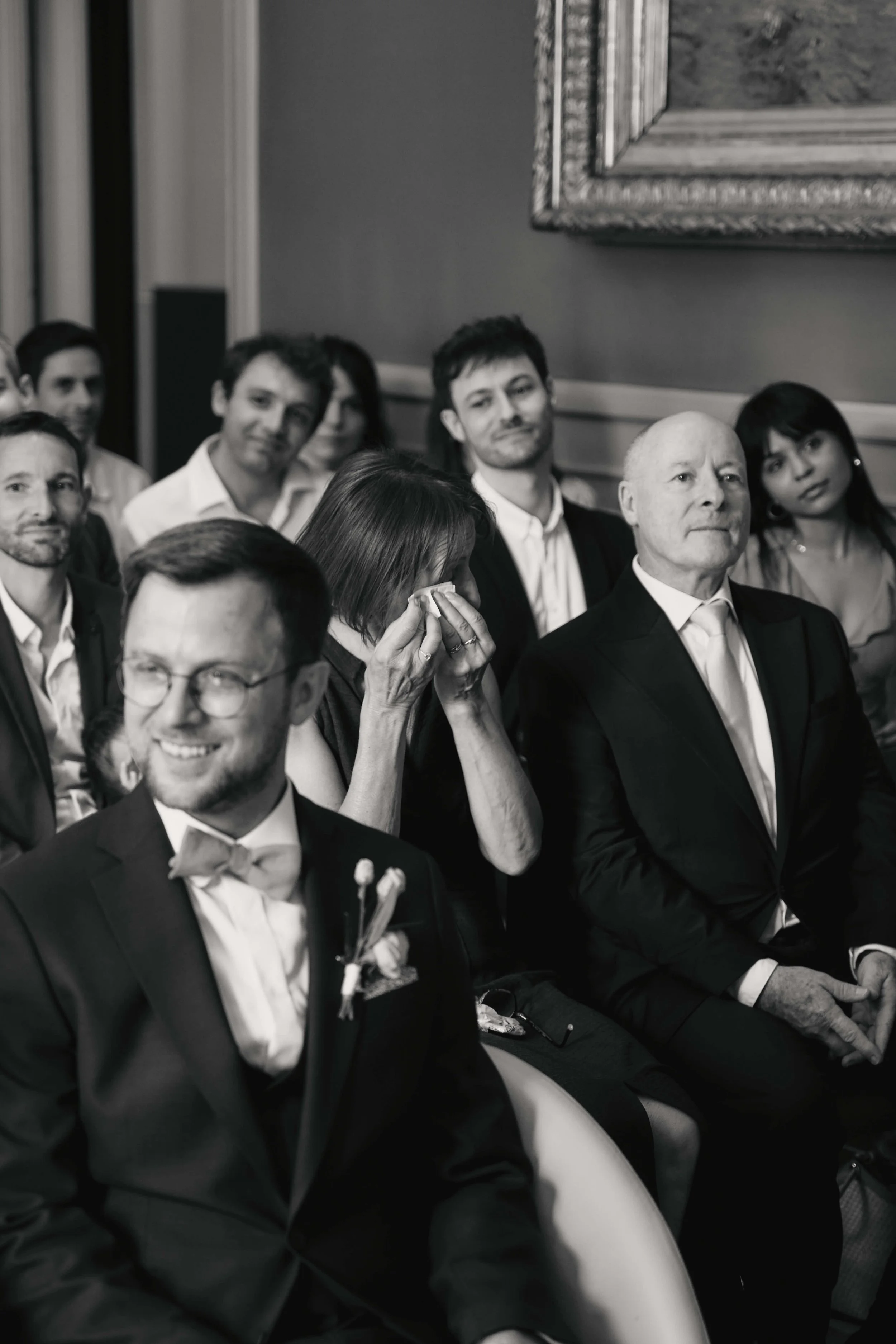 Group of people in formal attire attending an event, sitting in rows, with some looking ahead and one person wiping tears from her eyes.