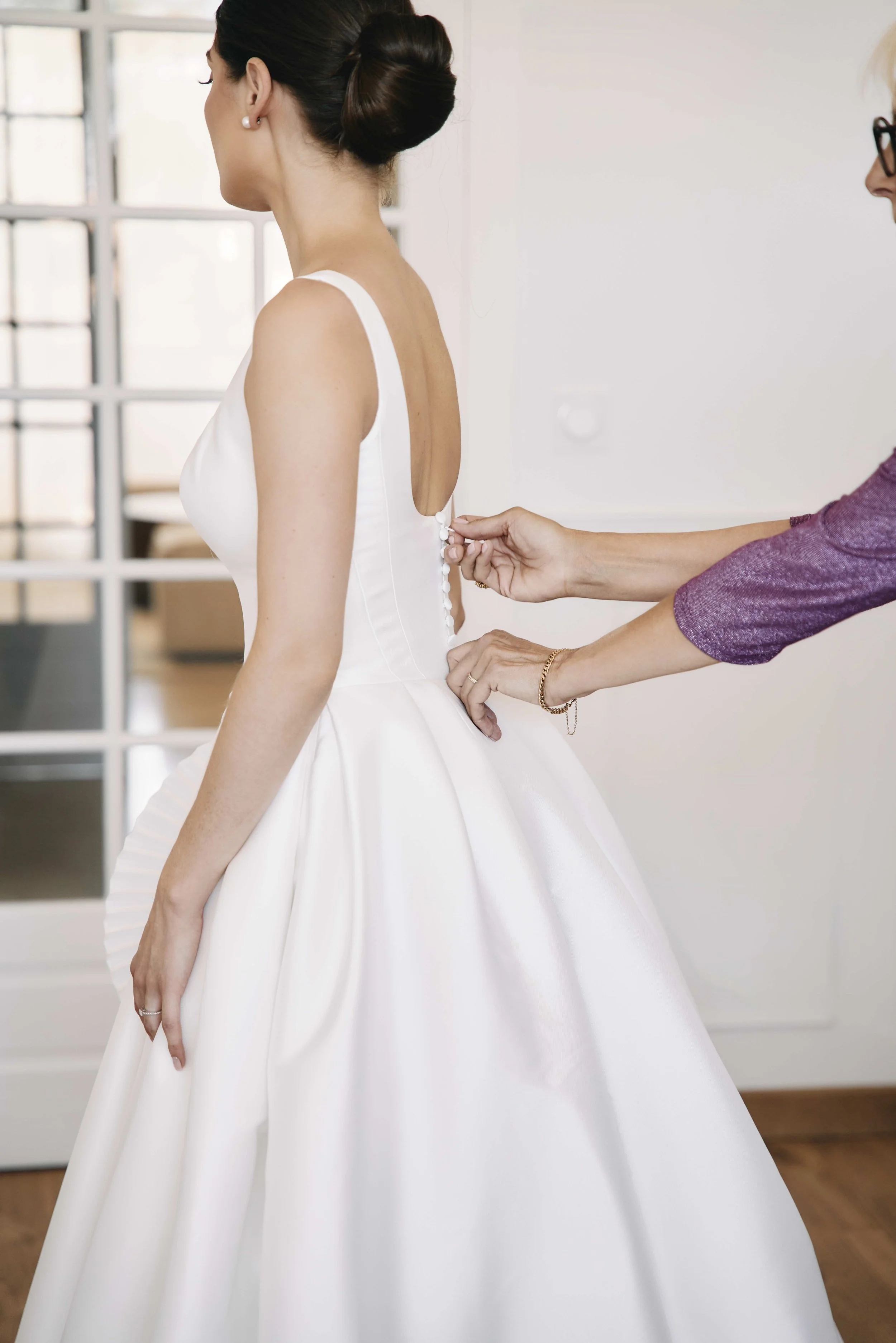 A bride in a white dress with a low back is being buttoned into her dress by a woman. The bride has dark hair styled in an elegant updo and is wearing pearl earrings. The background features a brightly lit room with large windows.