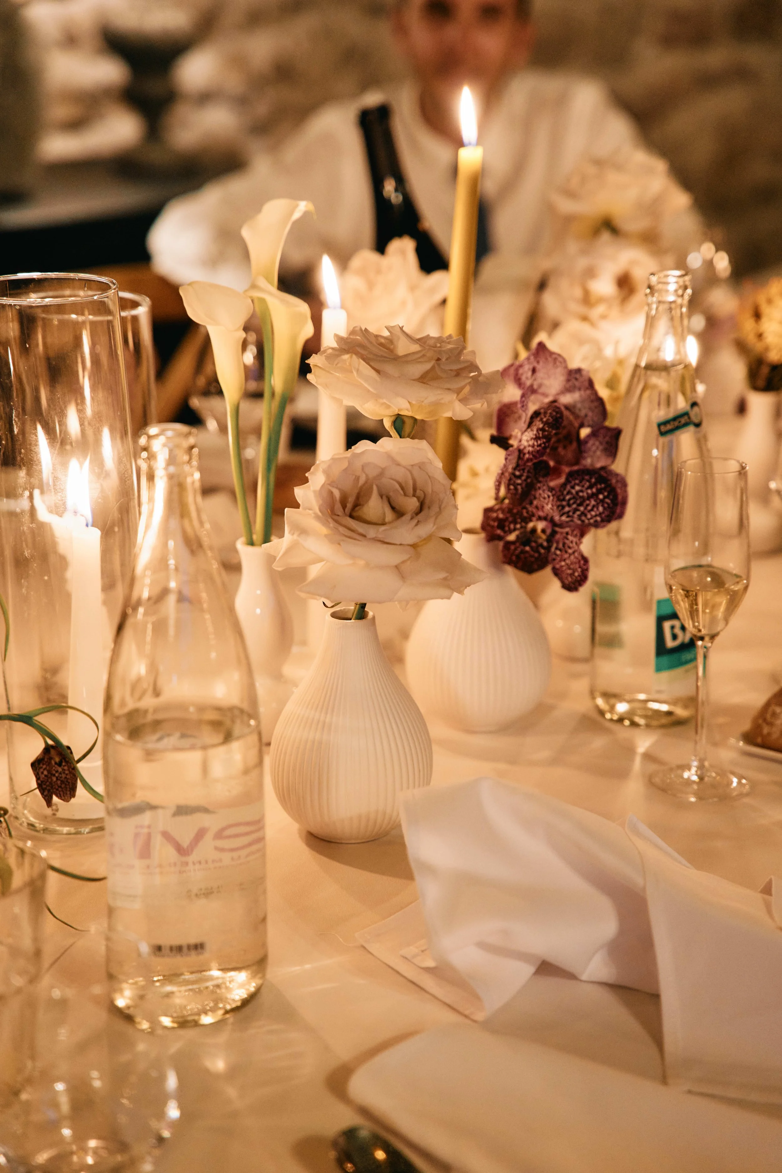 A dining table with vases of white calla lilies and orchids, lit candles, water bottles, glasses of wine, and a beige napkin on a white cloth tablecloth, with a blurred woman in the background.