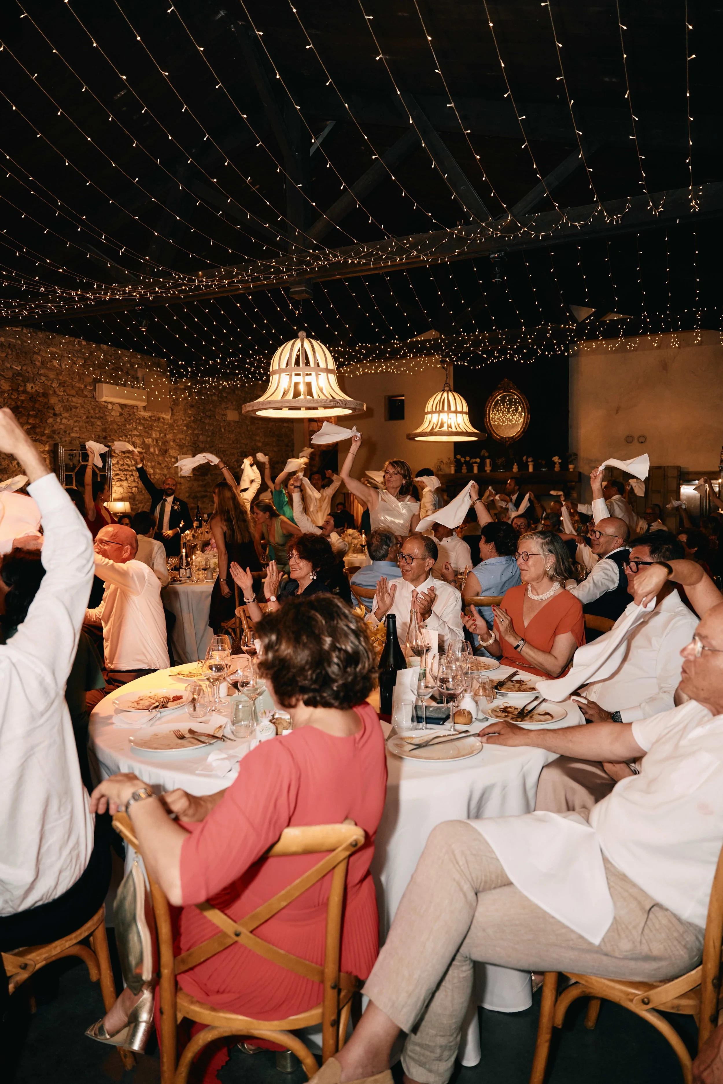 People celebrating at a dinner party with string lights hanging from the ceiling, some standing and dancing, others sitting at tables enjoying their meal.