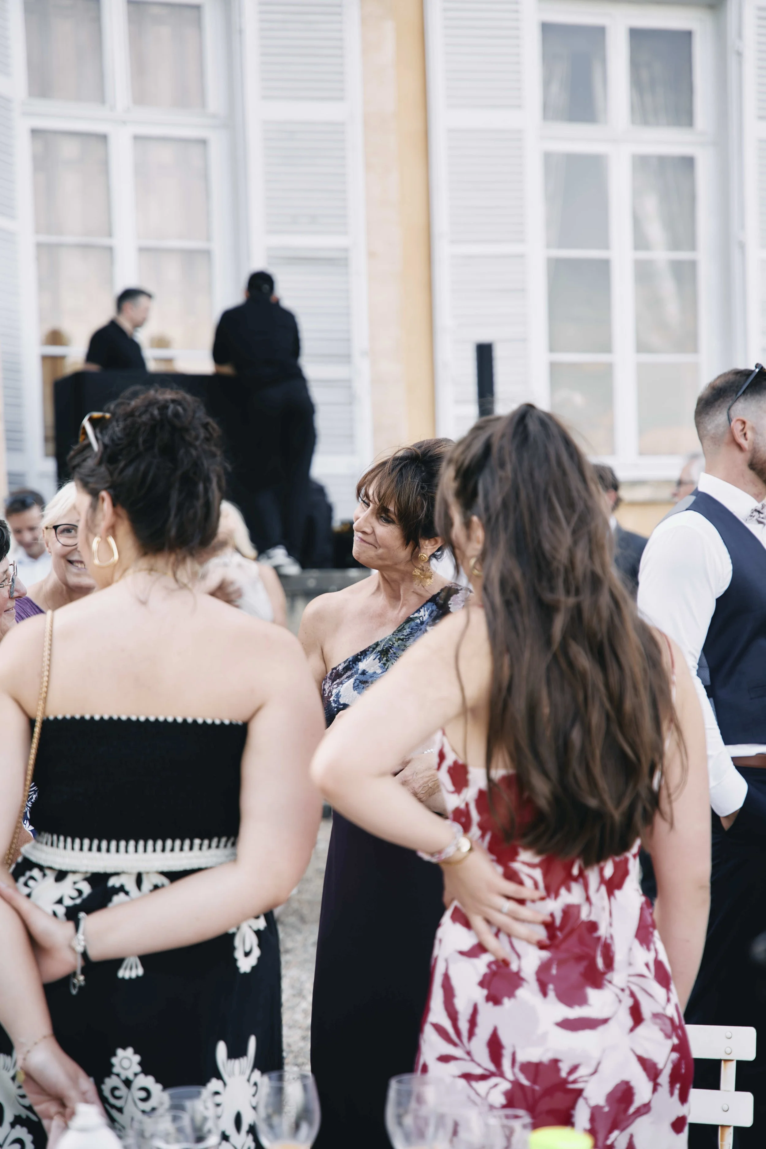 A group of people gathered outdoors at an event, dressed in semi-formal or formal attire, with some engaged in conversation. In the background, there are large windows and two individuals standing near a table, with one possibly performing or speakin
