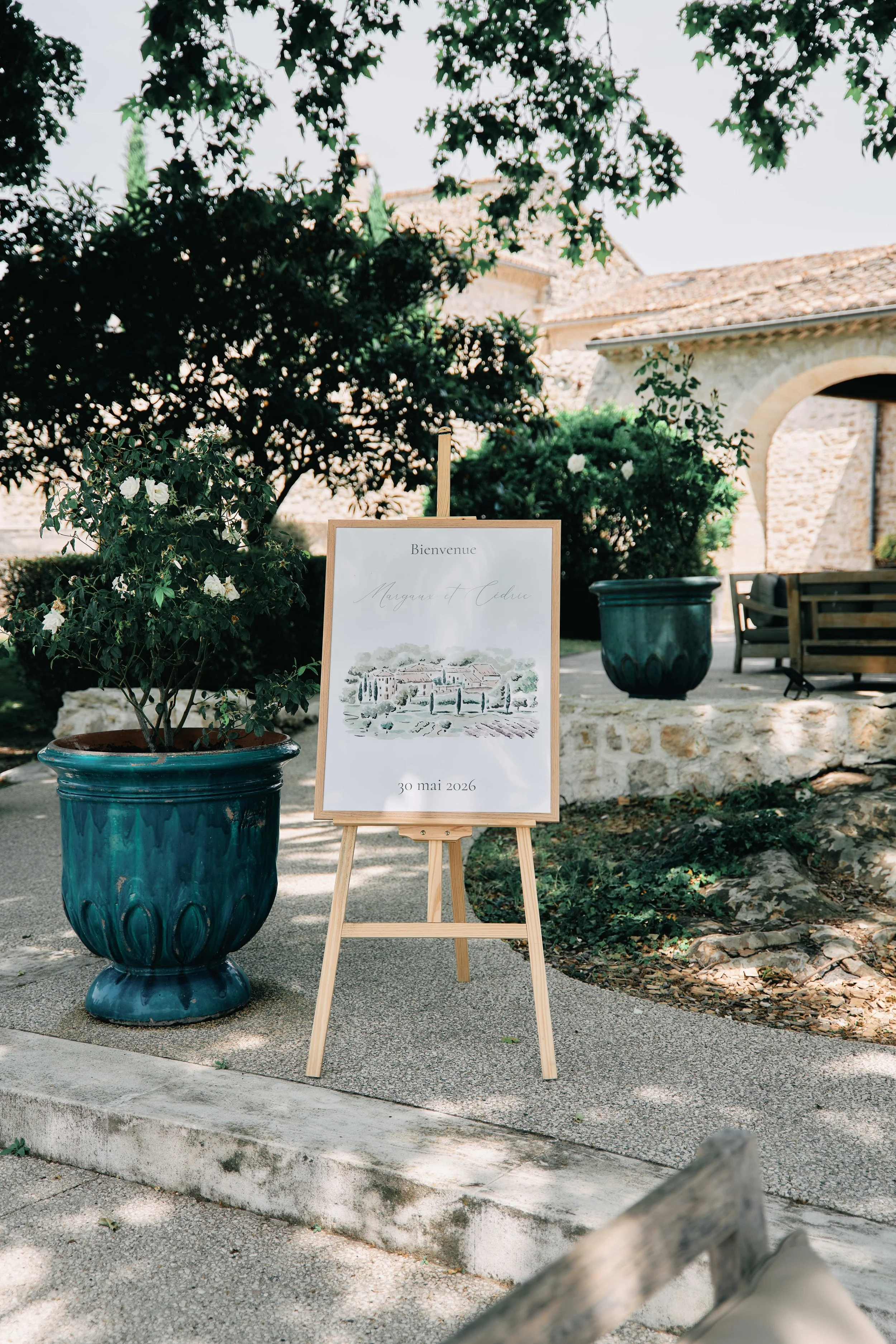 A welcome sign for Margaux and Cédric's wedding on May 30, 2026, is displayed on an easel on a sidewalk, surrounded by lush bushes and large flowerpots, with a rustic stone building in the background.
