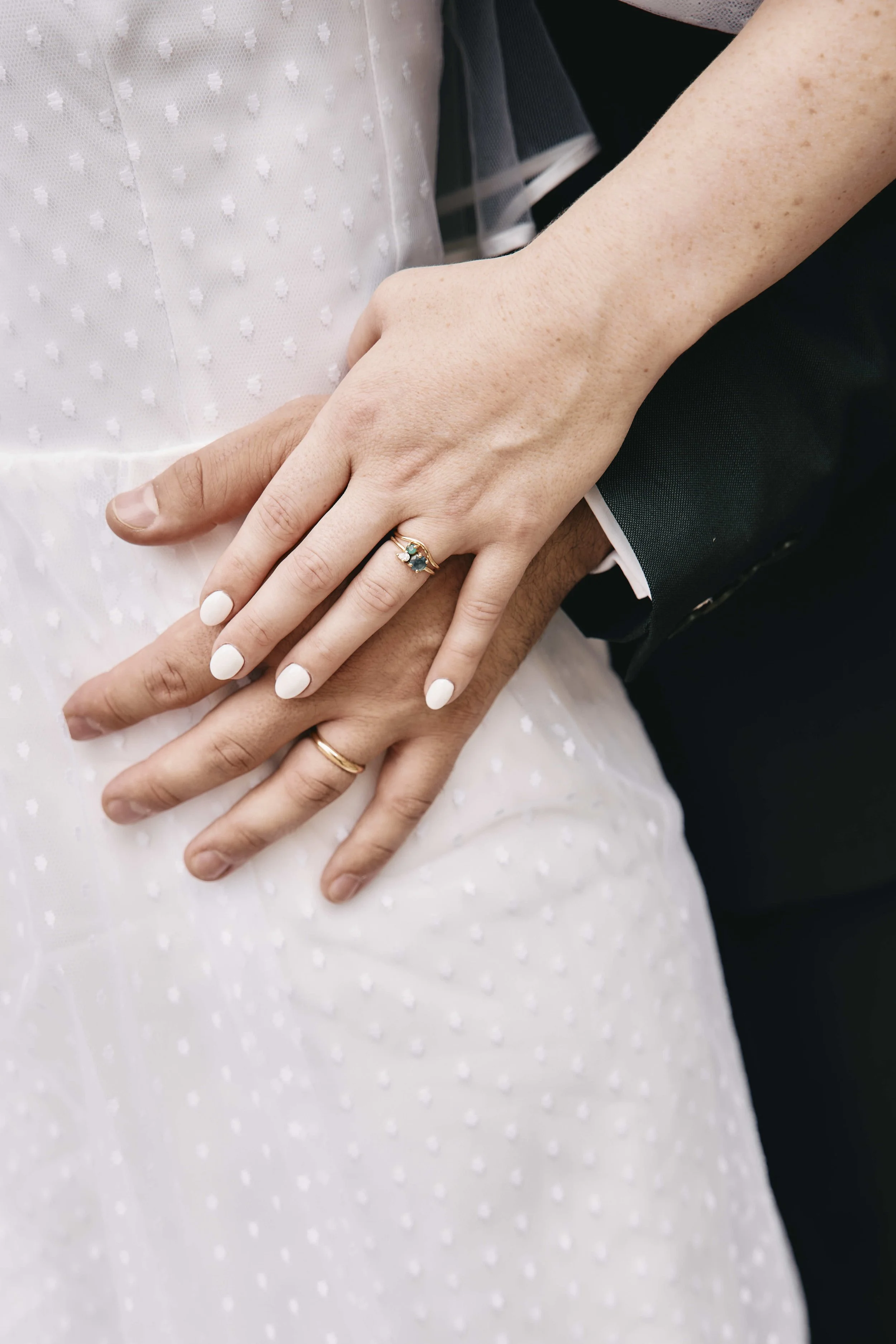 Close-up of a couple's hands intertwined, showing wedding rings, with the bride wearing a white dress and the groom wearing a dark suit.