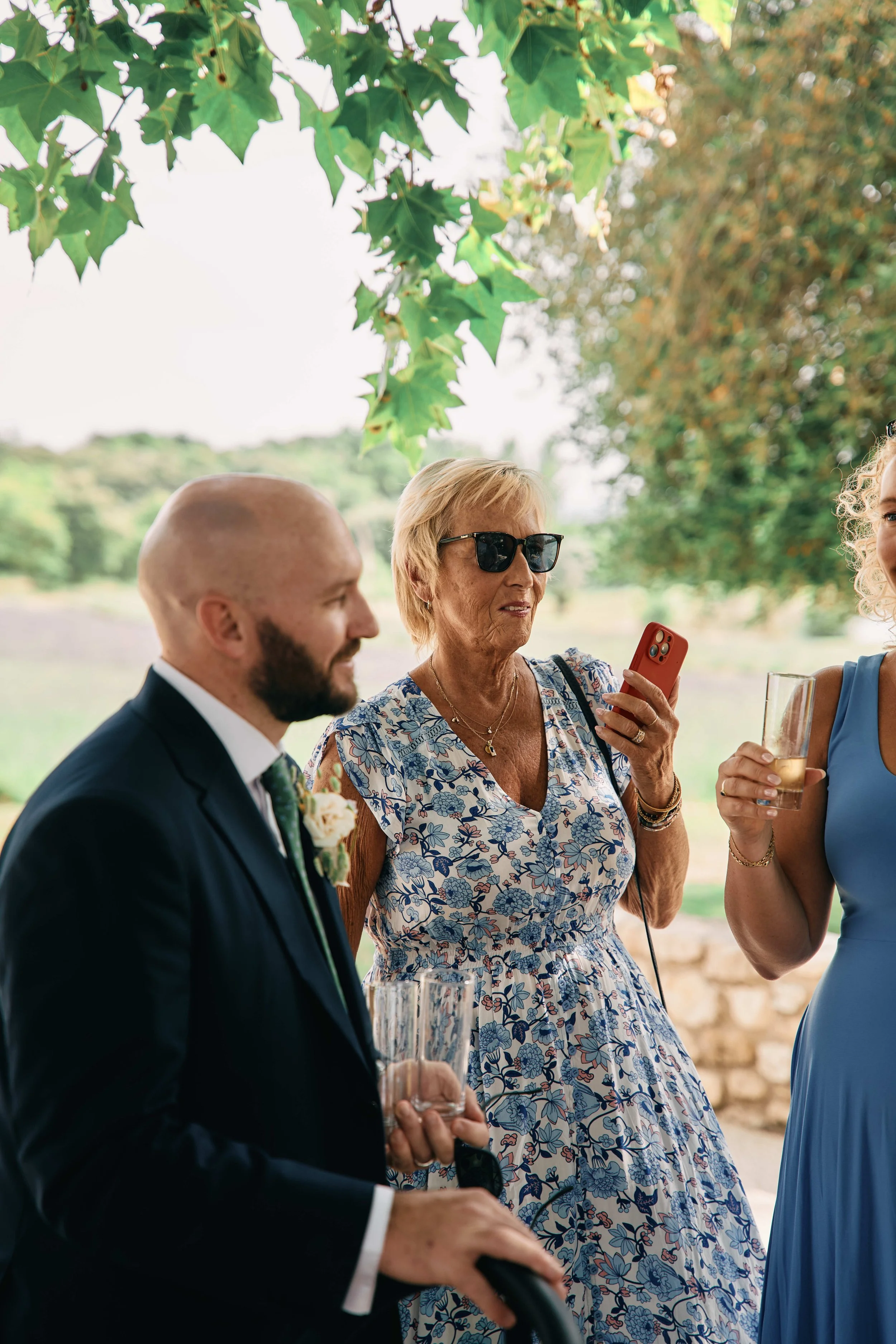 Group of people at an outdoor gathering, with a woman wearing sunglasses and holding a red phone as she takes a photo or video, and a man in a suit holding a drink, under a tree with green leaves.