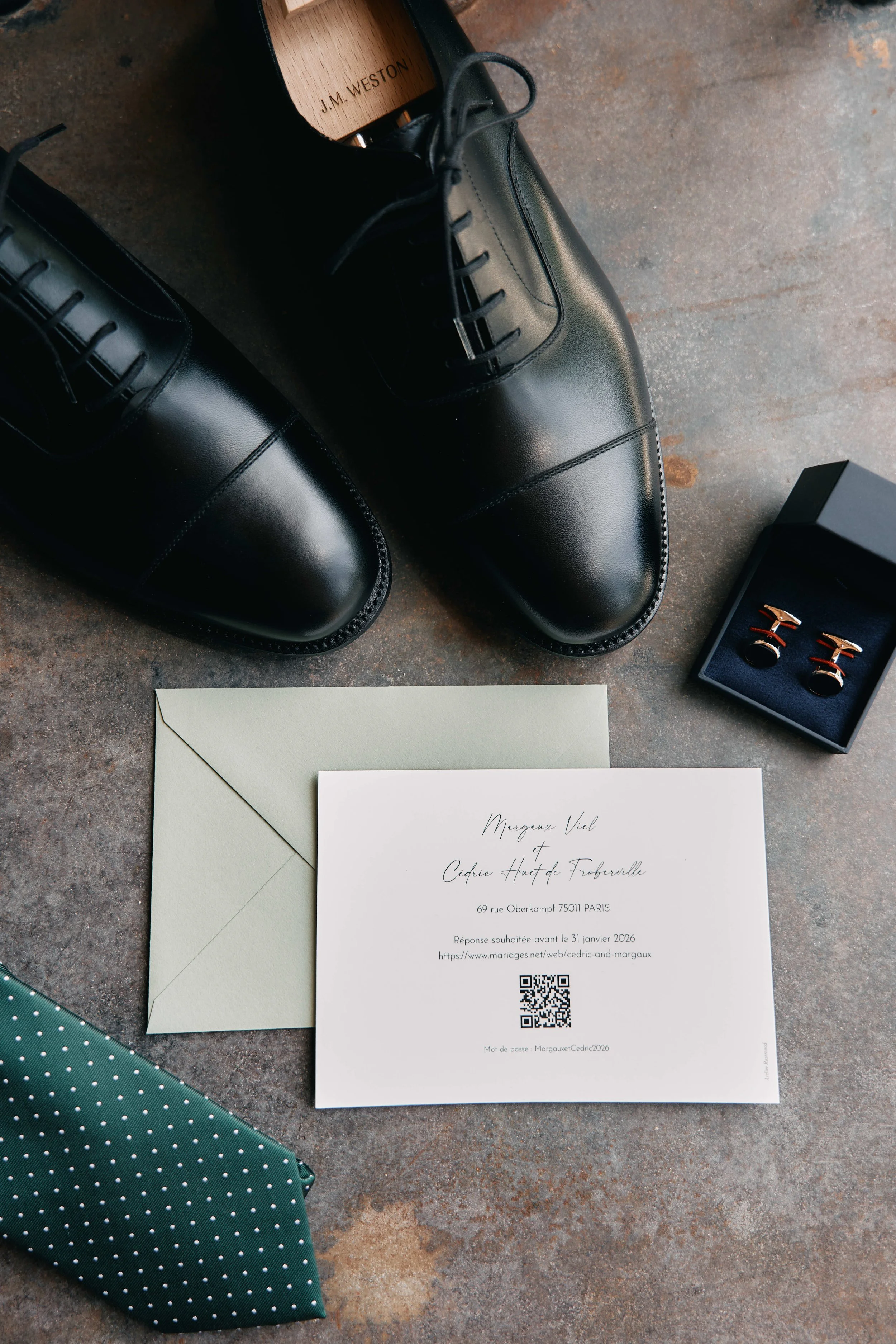 A pair of black leather dress shoes, a green polka dot tie, a white envelope, a white invitation card, a navy blue box with jewelry, and a wooden shoehorn.