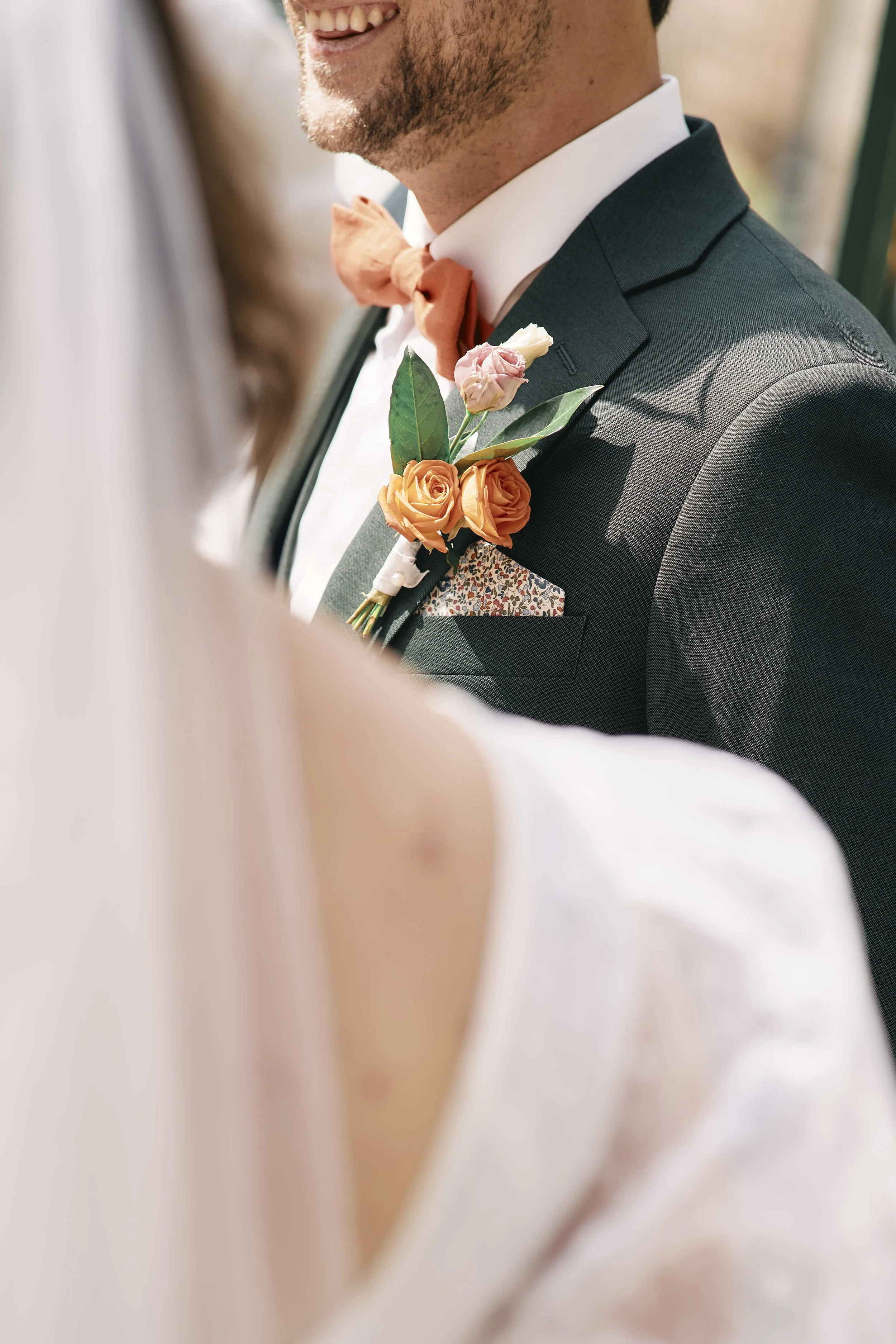 Close-up of a groom smiling, wearing a dark suit, white shirt, peach-colored bow tie, and a boutonniere with peach and white roses and green leaves.