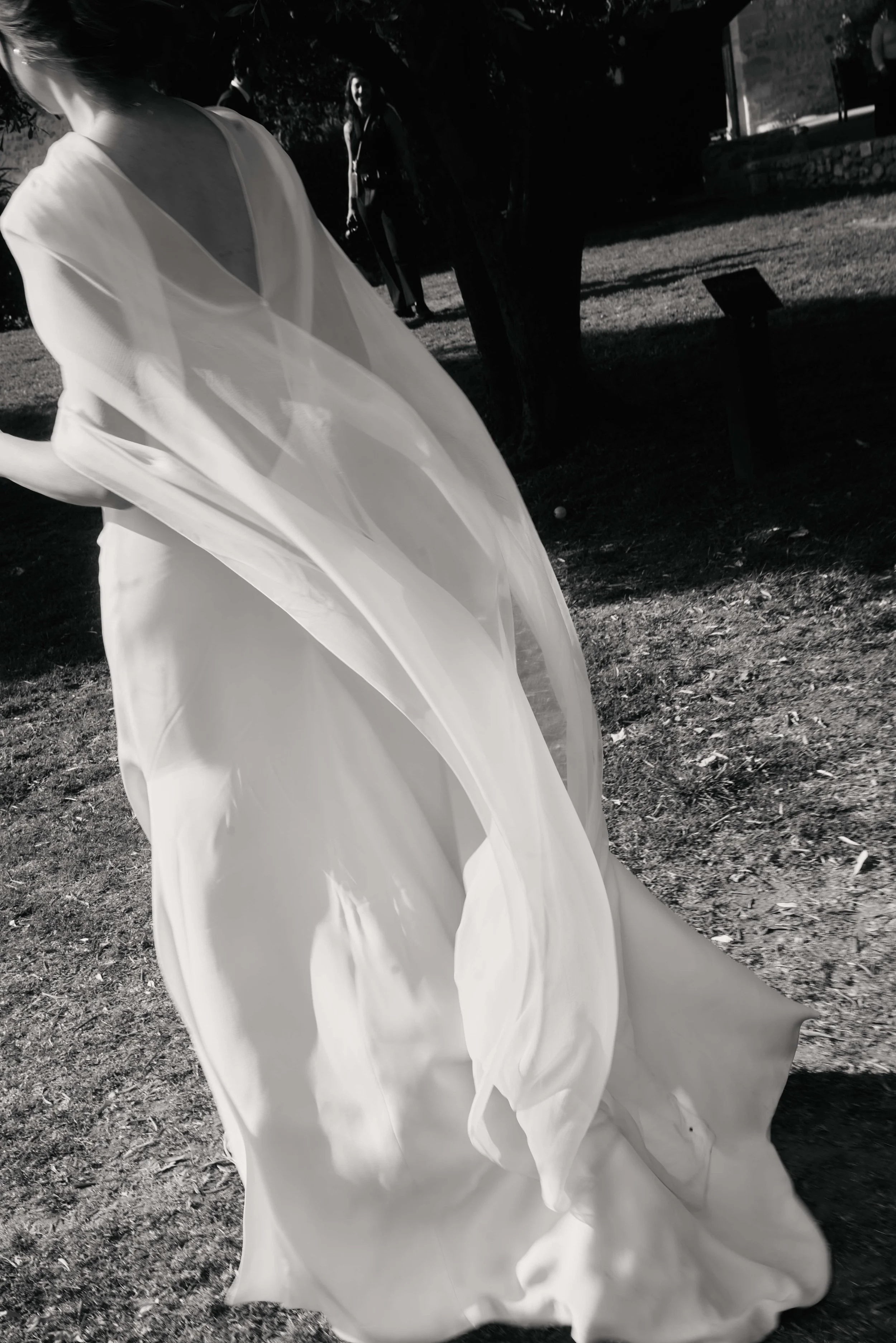 Woman in a flowing white dress outdoors with another person in the background.