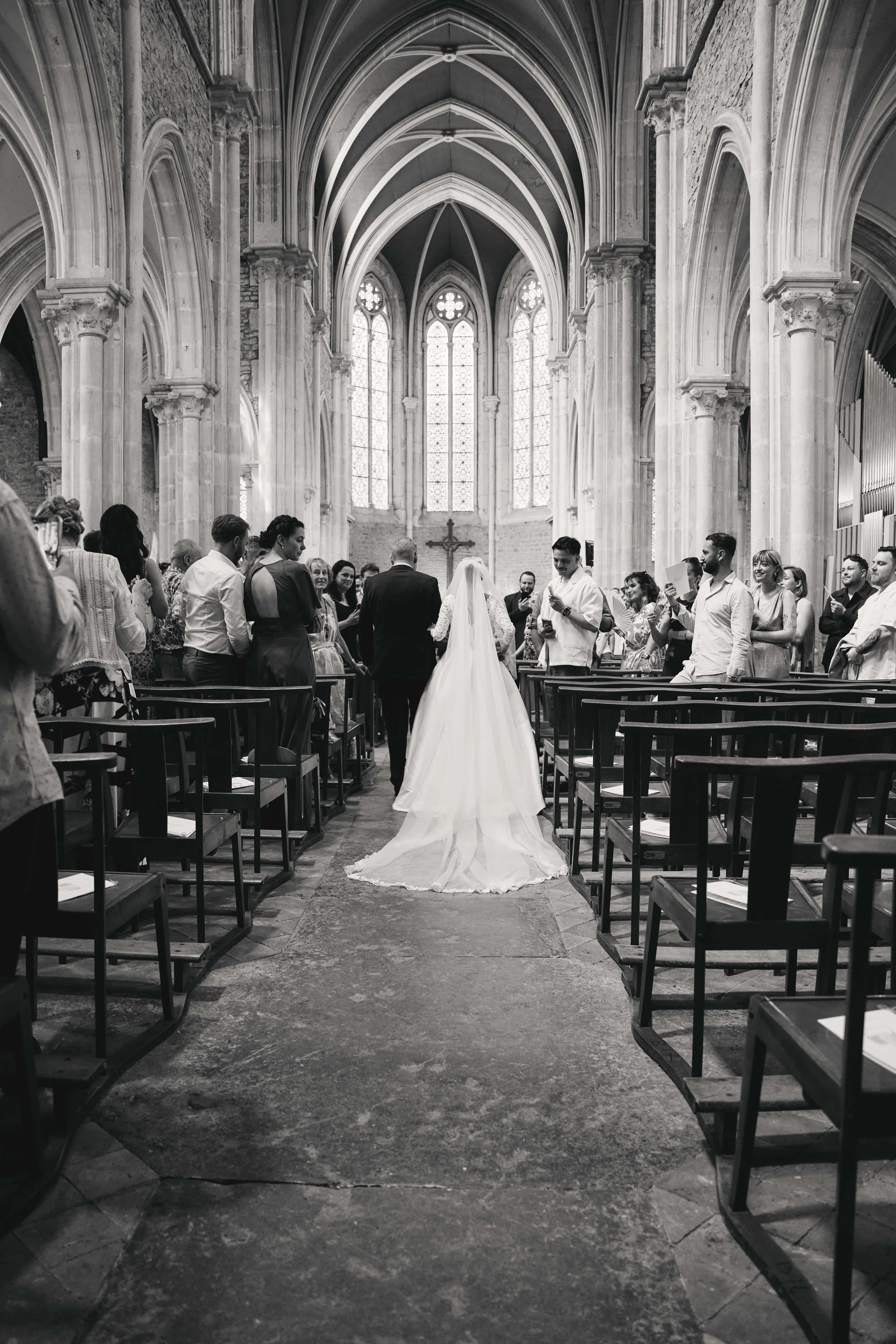 Black and white photo of a wedding ceremony inside a Gothic-style church with tall stained glass windows, stone arches, and pews. The bride and groom stand at the altar under a cross, surrounded by guests.