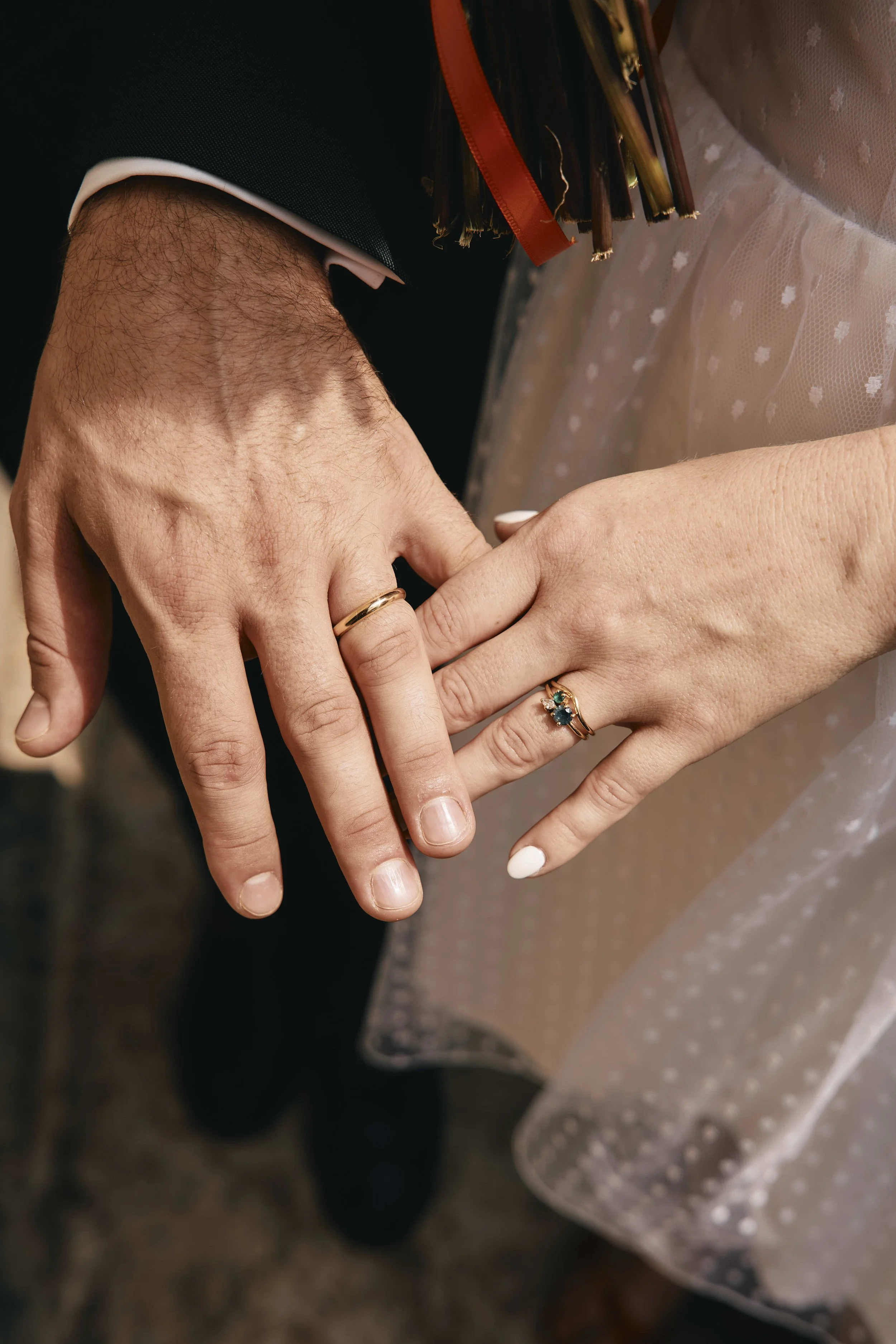 Close-up of a bride and groom's hands with wedding rings, holding each other, with wedding dresses visible in the background.