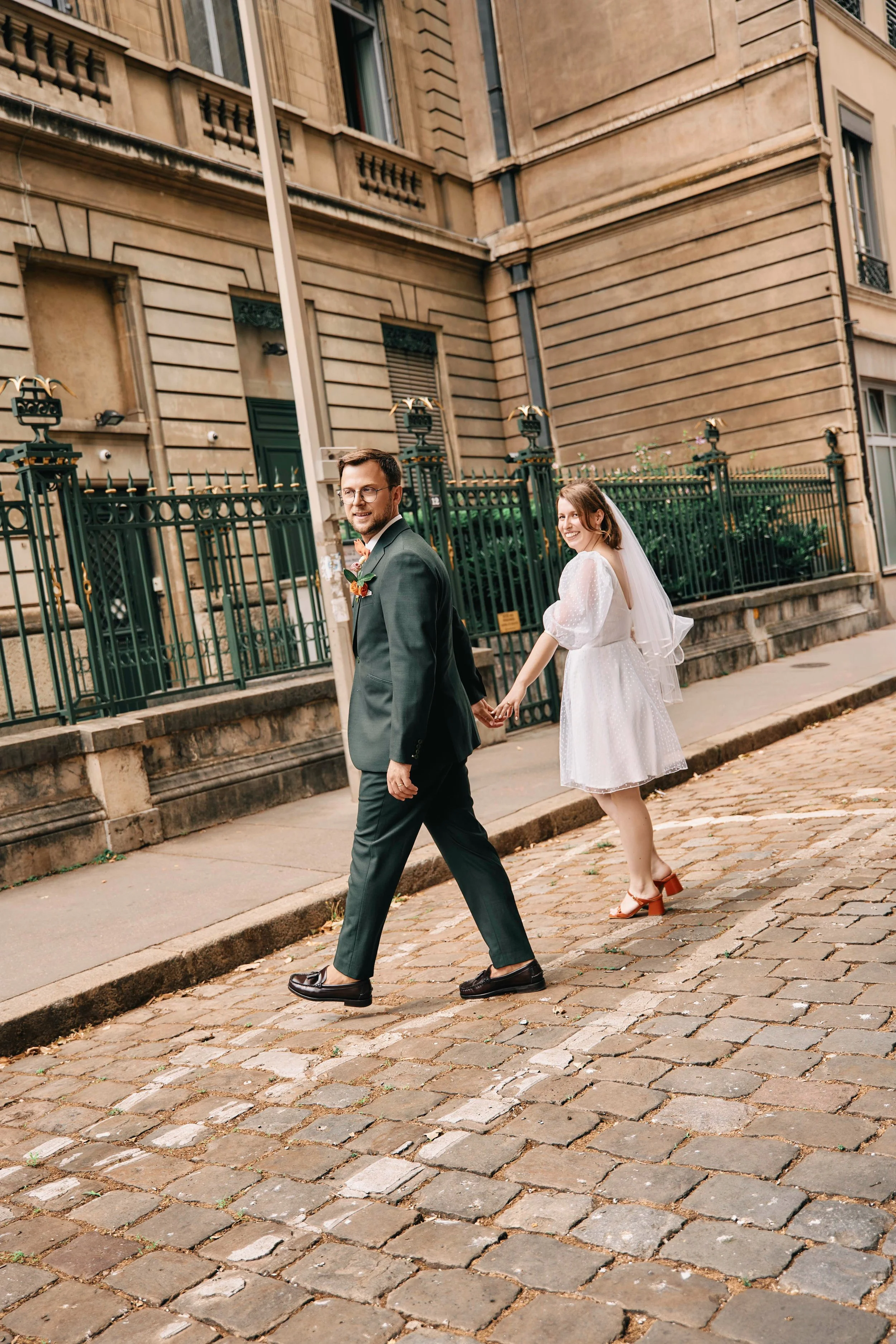 A smiling bride and groom walking hand in hand on a city sidewalk, with a historic building and black iron fence in the background.