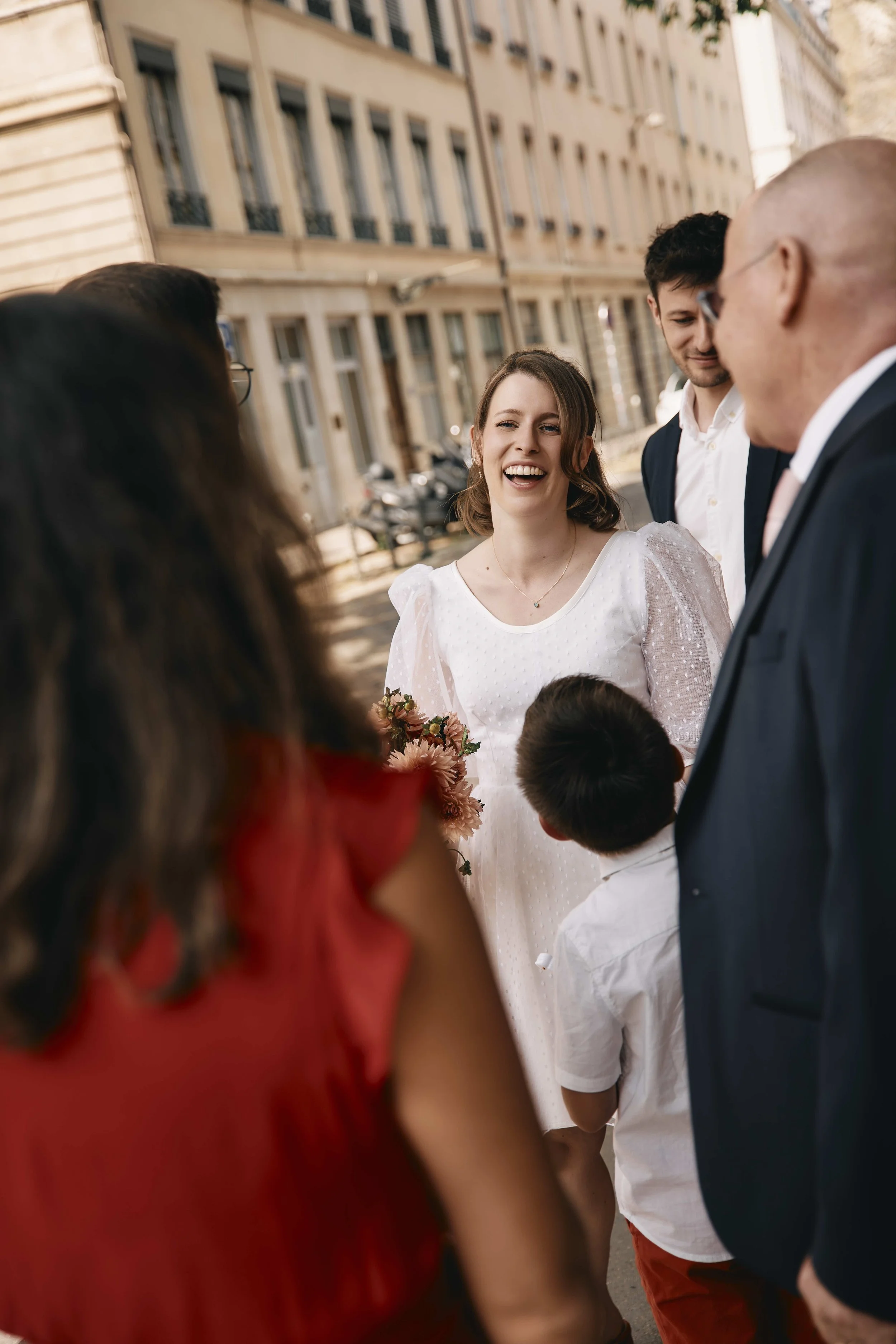 Four people chatting outdoors on a city street, children visible in the foreground, smiling woman in white dress in the center.
