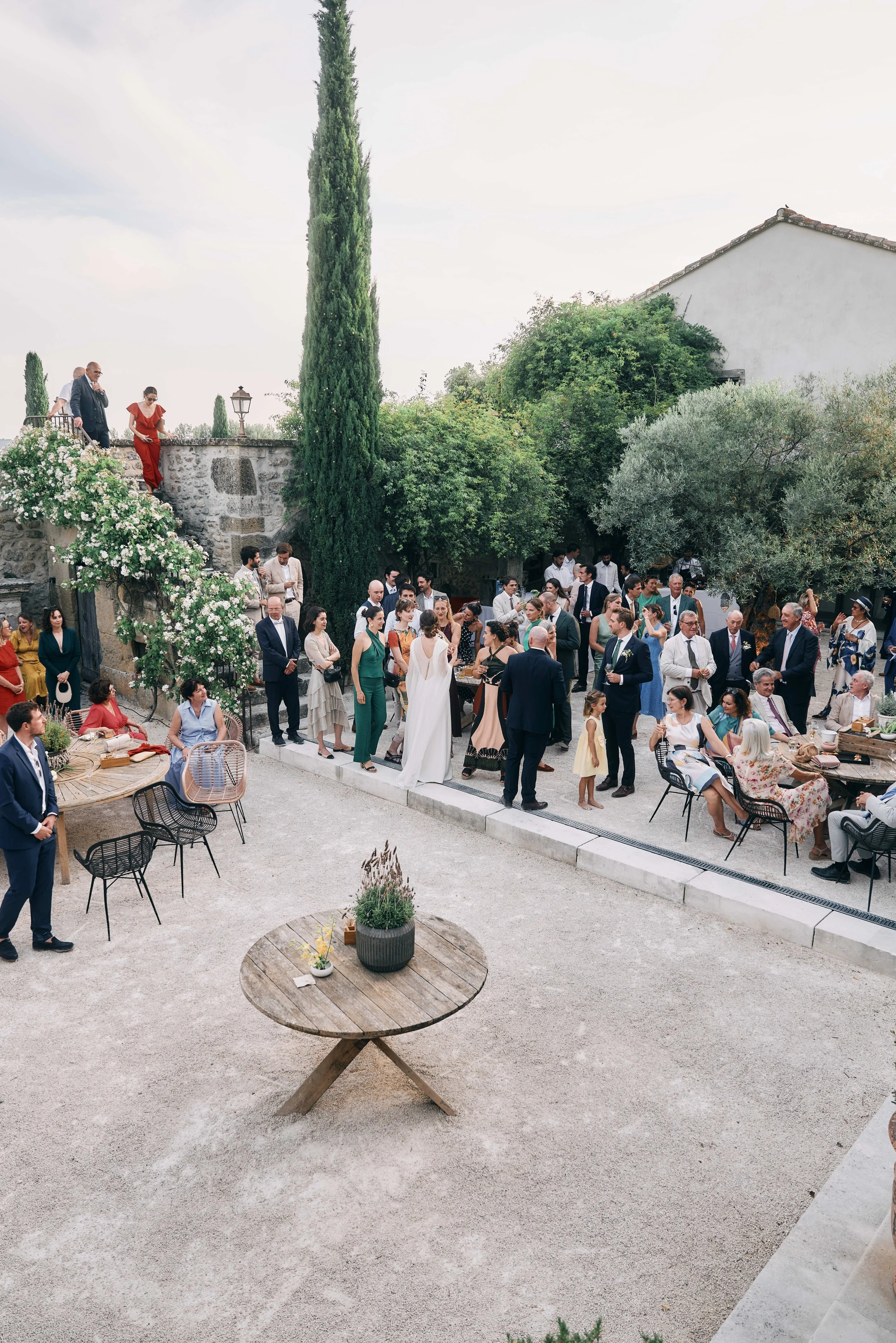 Outdoor gathering of people in formal attire at a garden with trees and plants, some standing on stone steps and others mingling around tables.