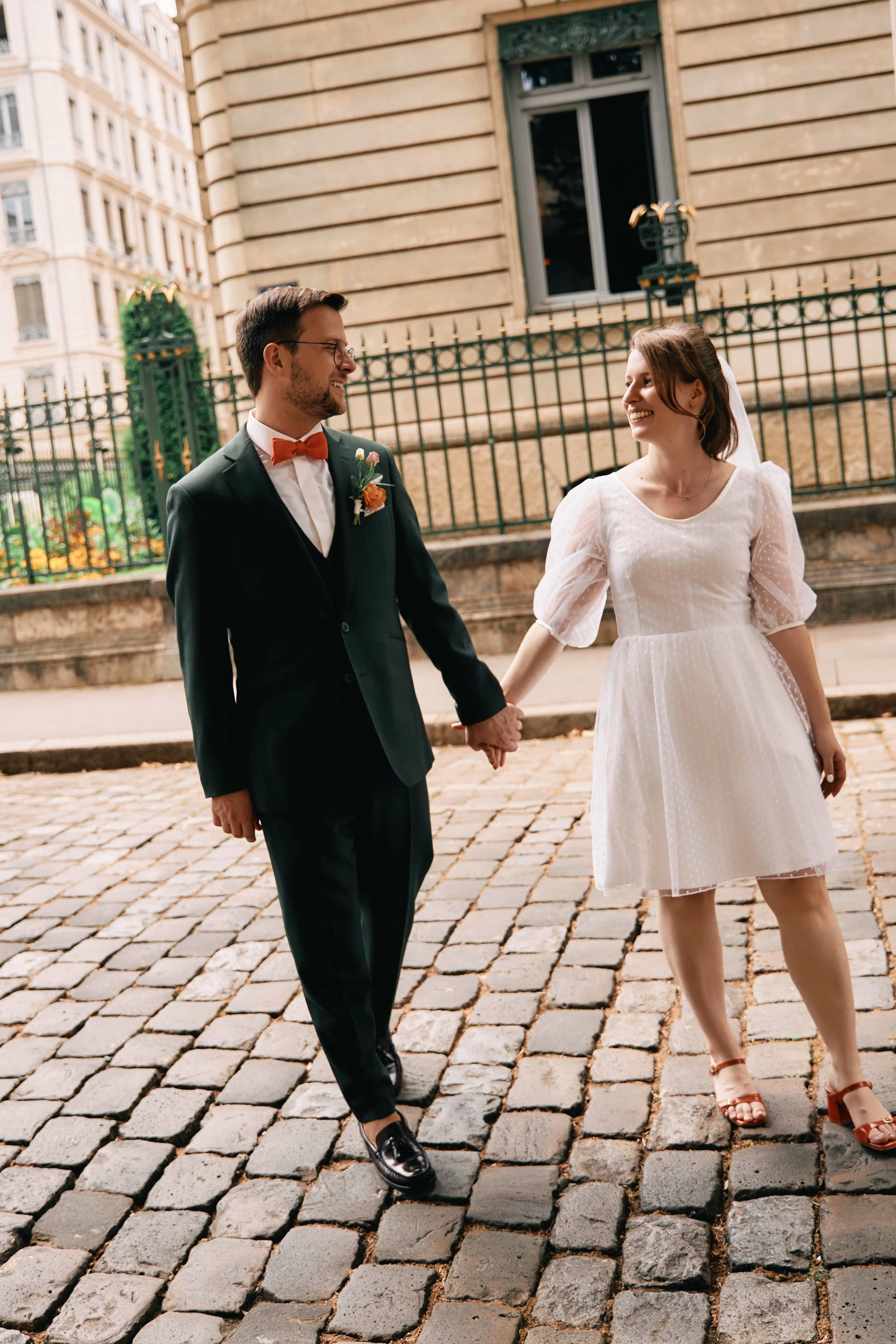 A man and woman holding hands and smiling at each other while walking on cobblestone street in front of a historic building with a black iron fence.