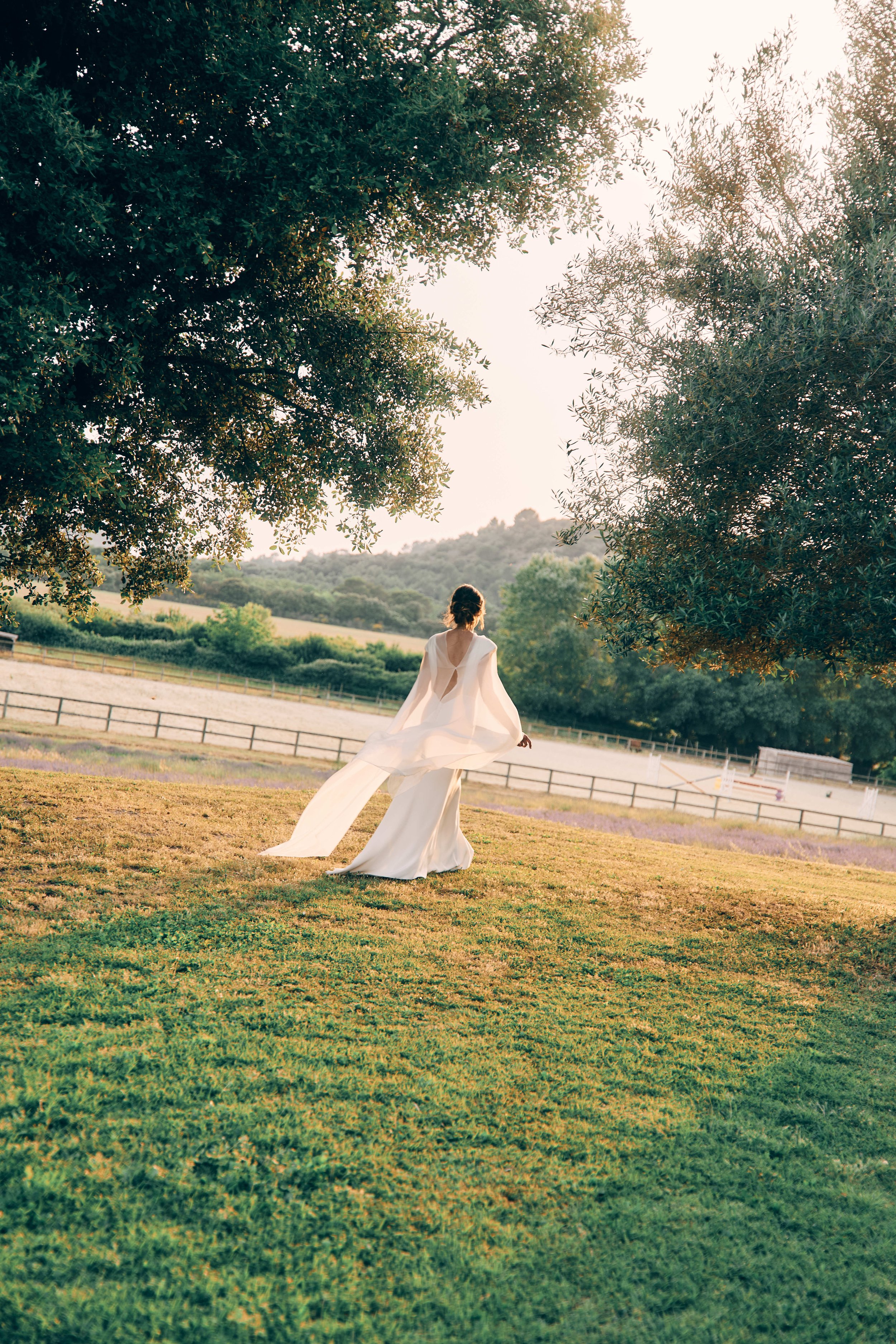 A woman in a long white dress walking on a grassy field with large trees and a distant hilly landscape during sunset.