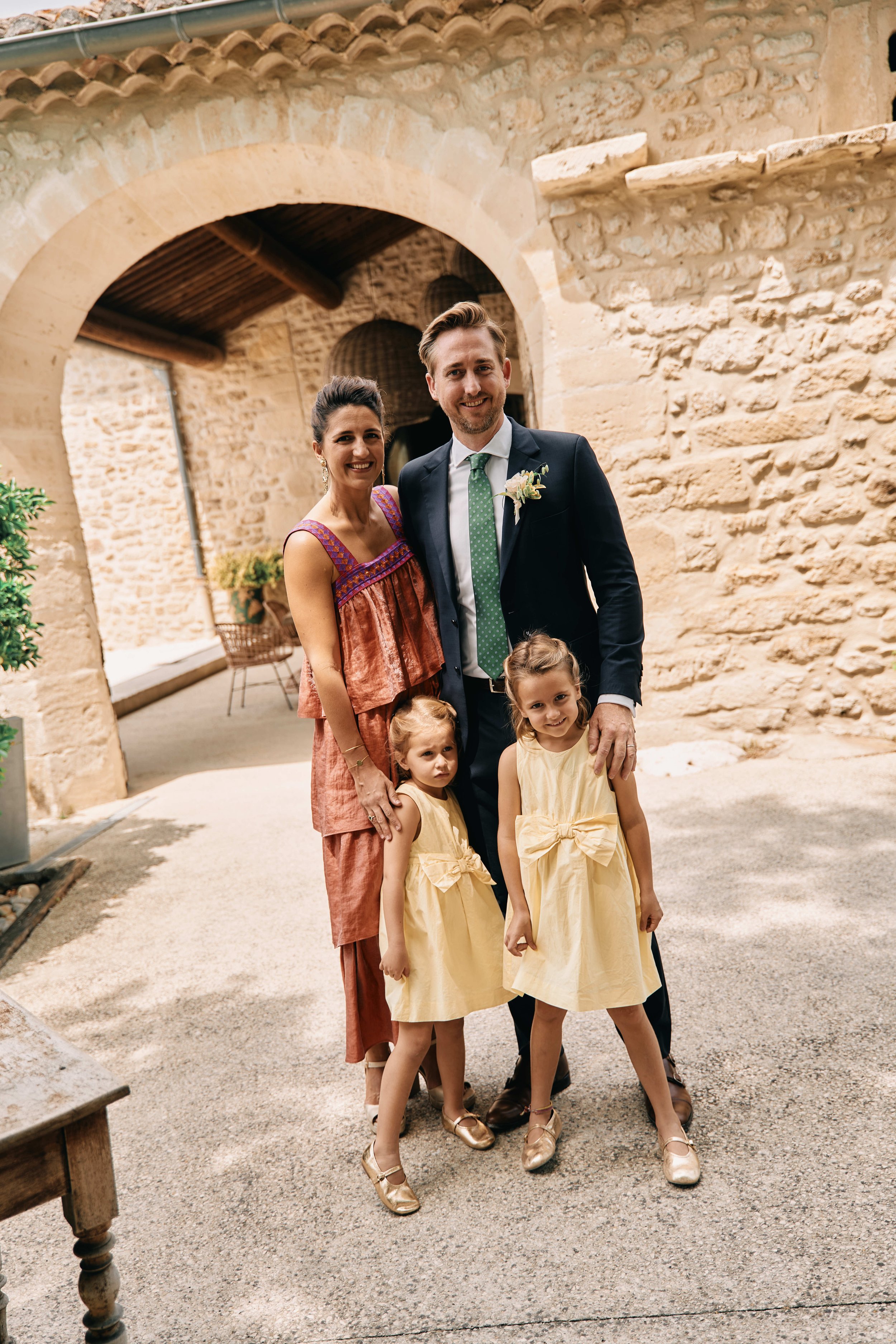 Four people, two adults and two children, are standing together outdoors in a courtyard with stone walls. The adults are dressed formally, with the man in a suit and the woman in a colorful dress. The children are wearing matching yellow dresses. Eve