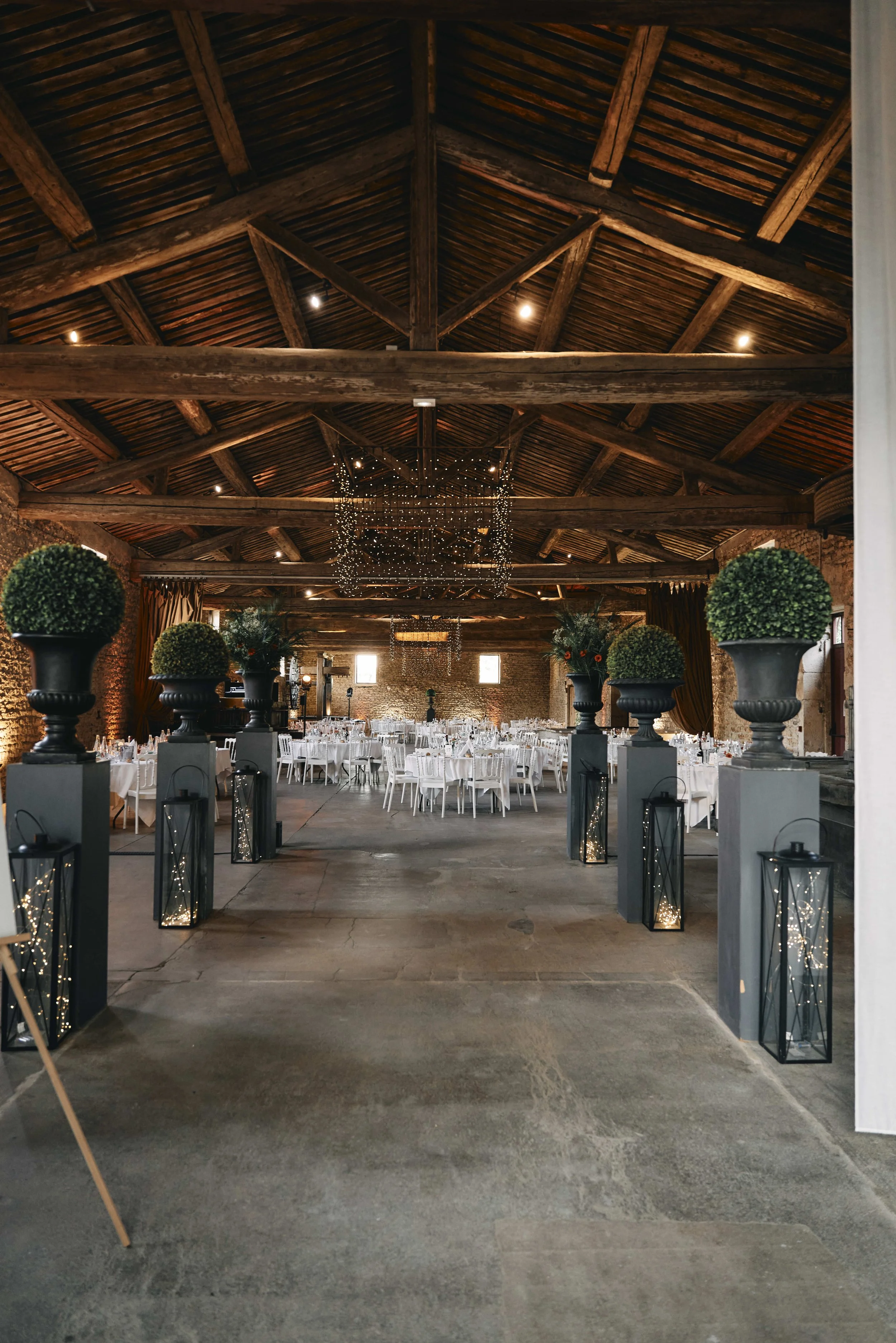 Interior of a rustic event hall with a high wooden ceiling, string lights, and tables set for a celebration, with decorative greenery and lanterns along the entrance pathway.