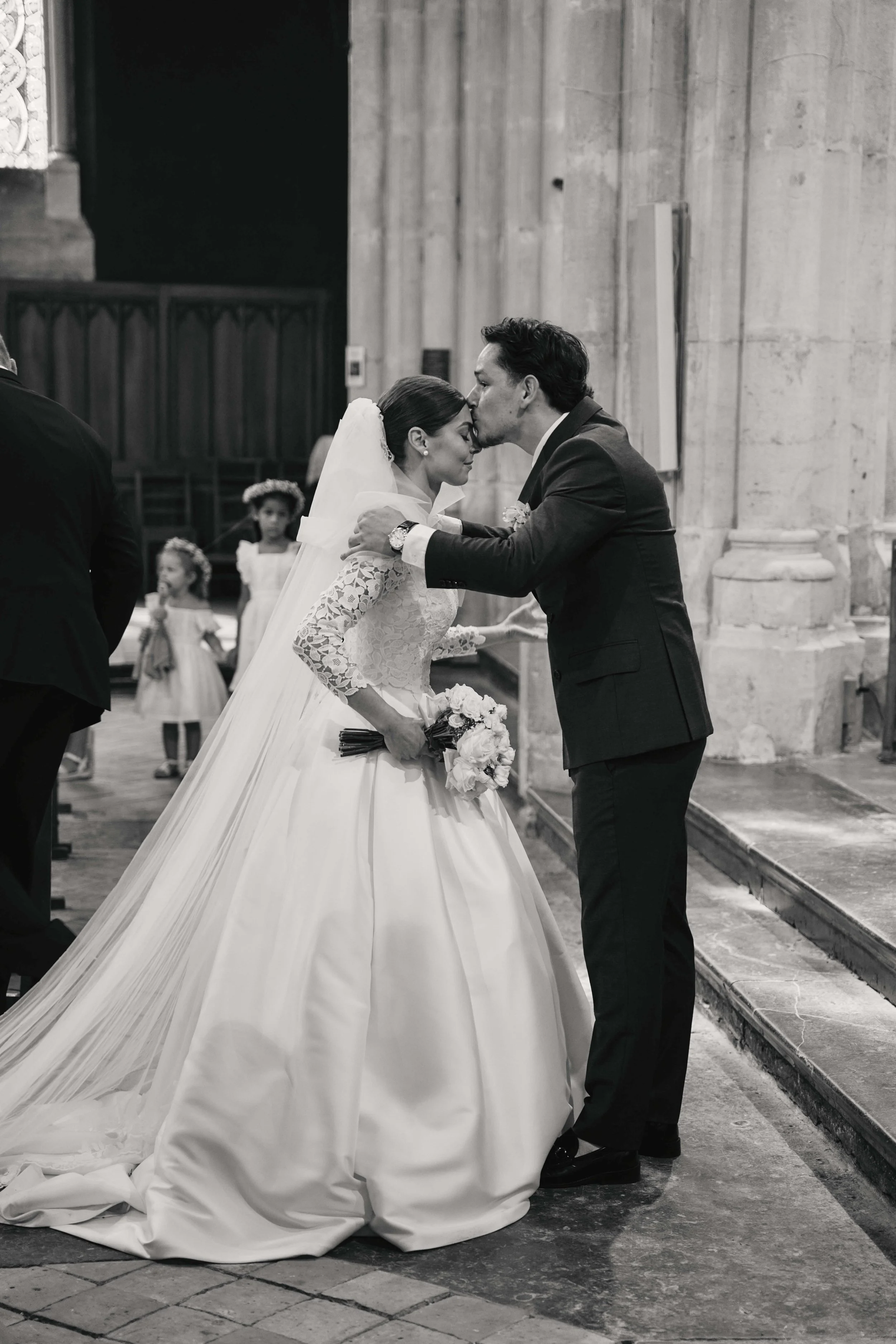 A black-and-white photo of a bride and groom kissing inside a church, with the bride holding a bouquet and wearing a wedding dress, and the groom dressed in a suit. Two young flower girls are in the background.