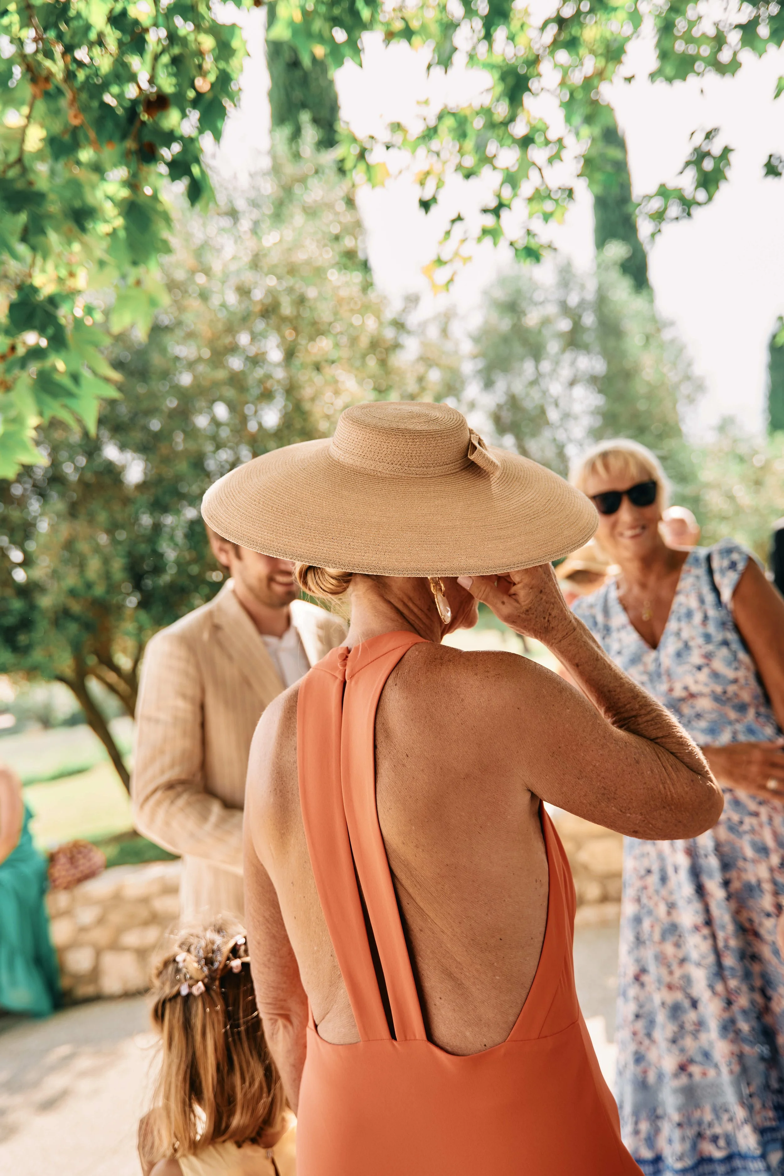 A woman wearing a wide-brimmed hat and a backless orange dress with a ribbon detail, standing outdoors among people at a social gathering in a park-like setting with trees and greenery.