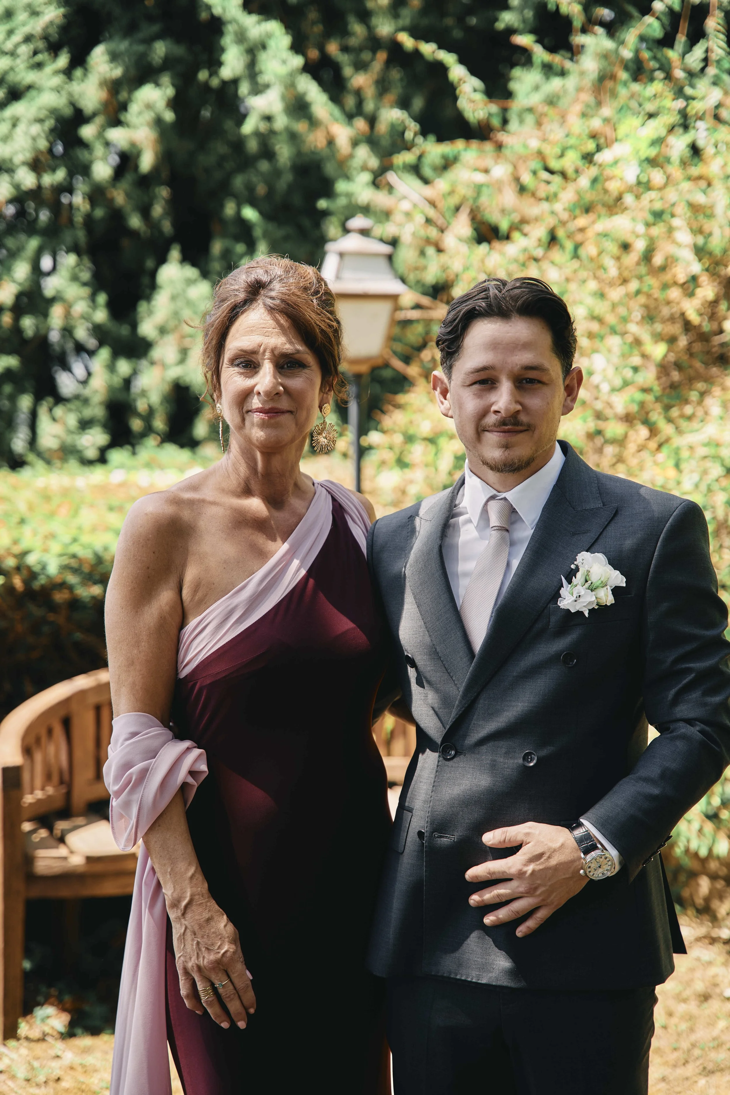 A middle-aged woman and a young man dressed in formal attire standing outdoors with greenery in the background.