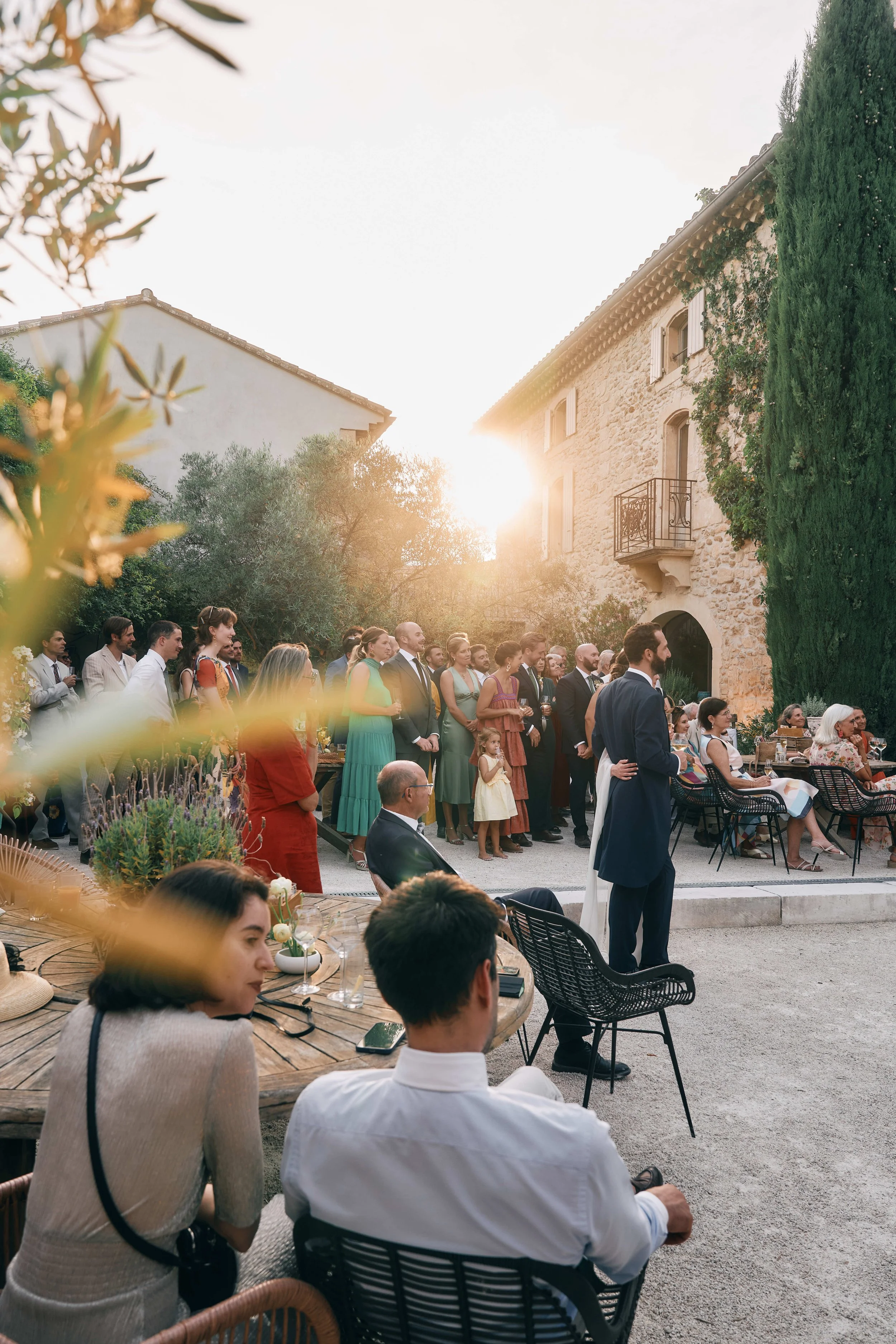 People dressed in formal attire gather outdoors at sunset, attending a wedding or reception in a rustic setting with stone buildings and lush greenery.