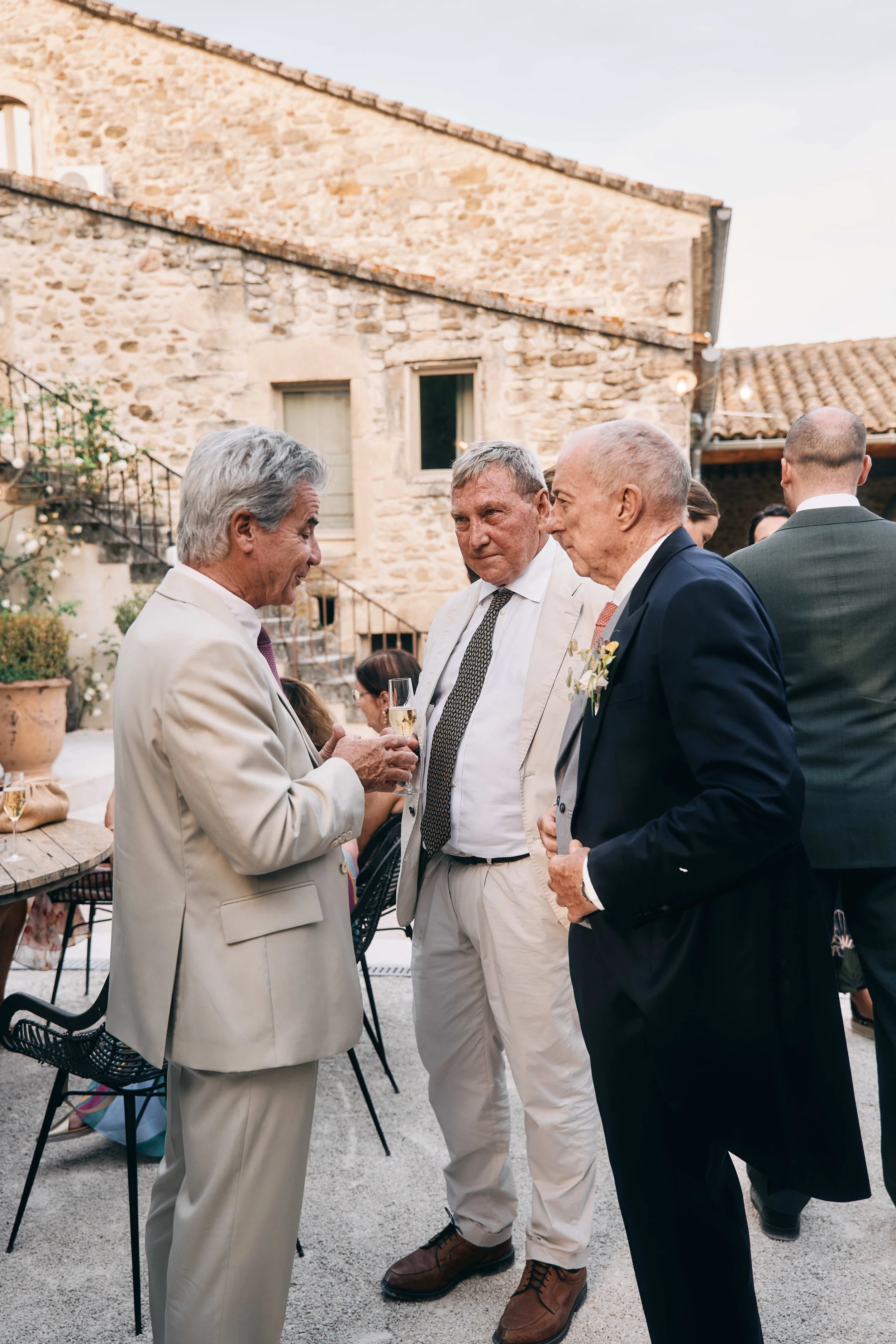 At a wedding reception, elderly men in suits are engaged in conversation outdoors in front of a stone building with a tiled roof. One man holds a glass of champagne, and others are listening or talking.