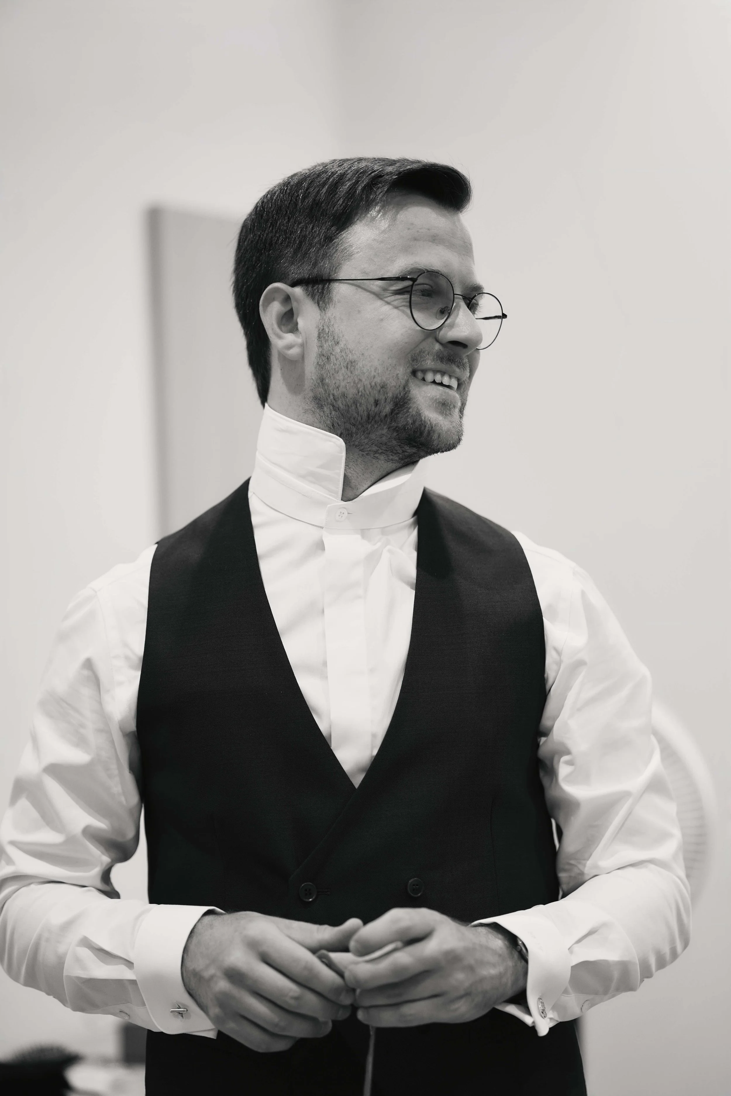 A man with glasses and dark hair, dressed in a white shirt and a dark vest, standing against a plain light background.