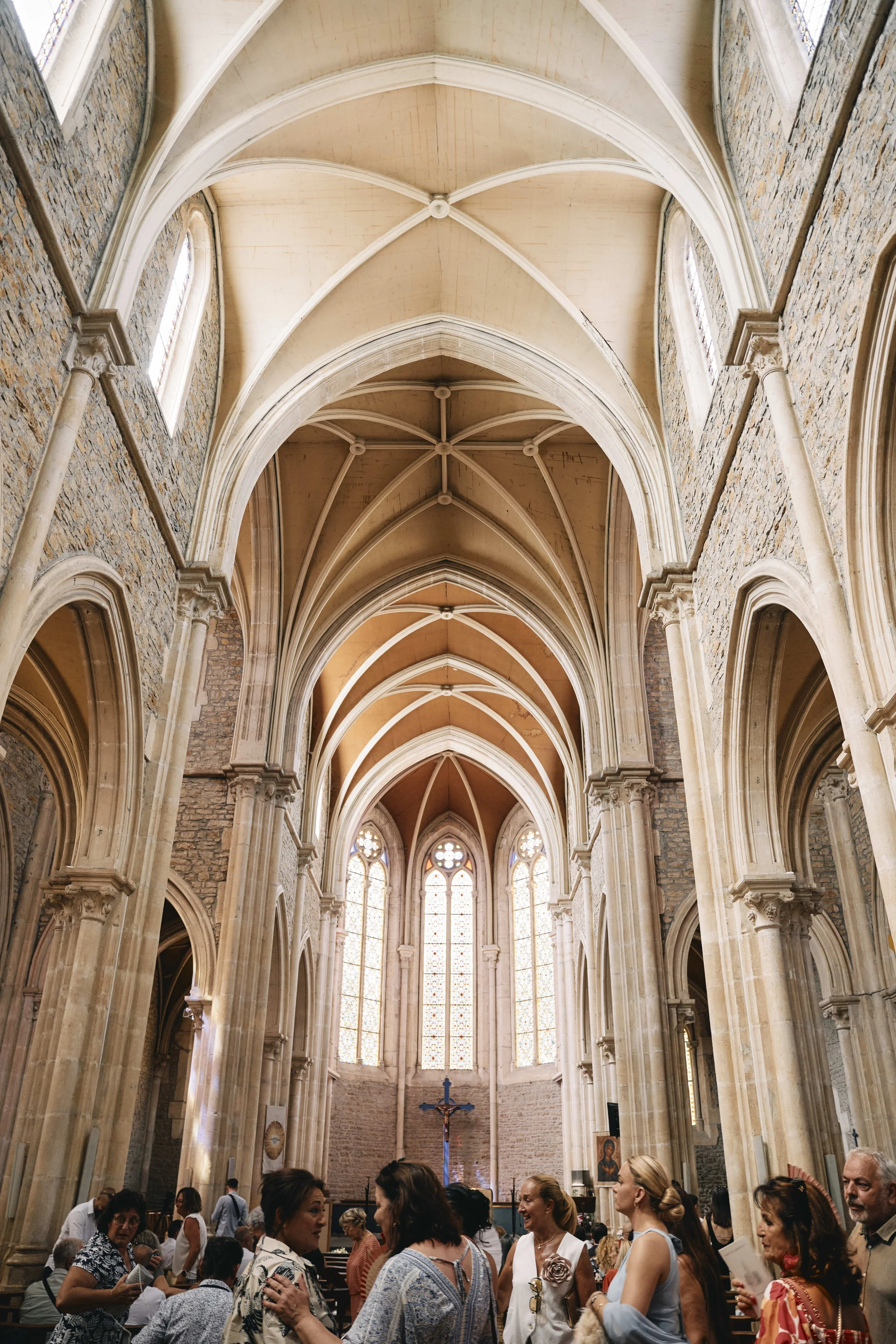 Inside a church congregation gathering with high gothic ceilings, stone walls, tall stained glass windows, and a crucifix at the altar, people are standing and engaging in conversation.