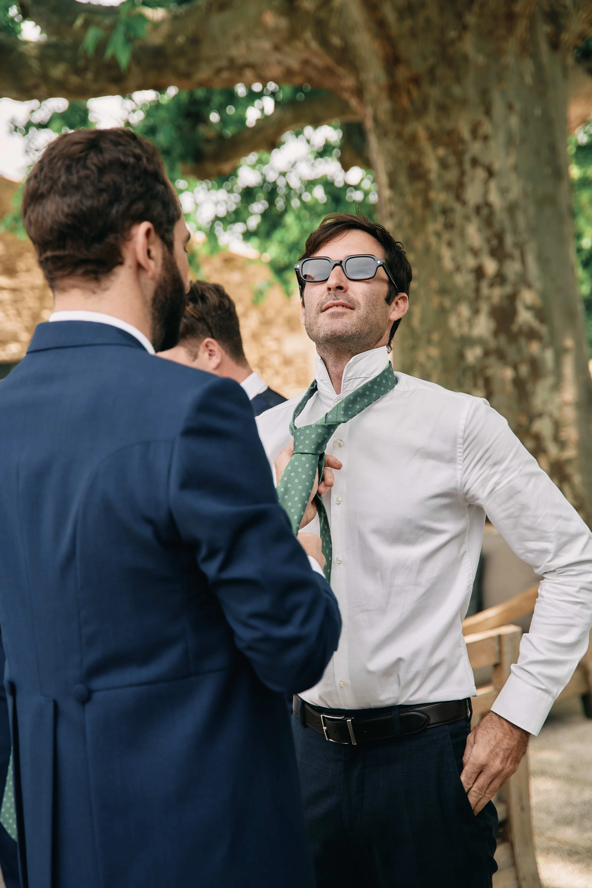 Three men in suits and sunglasses outdoors, one adjusting the tie of another, with large tree and wooden fence in background.