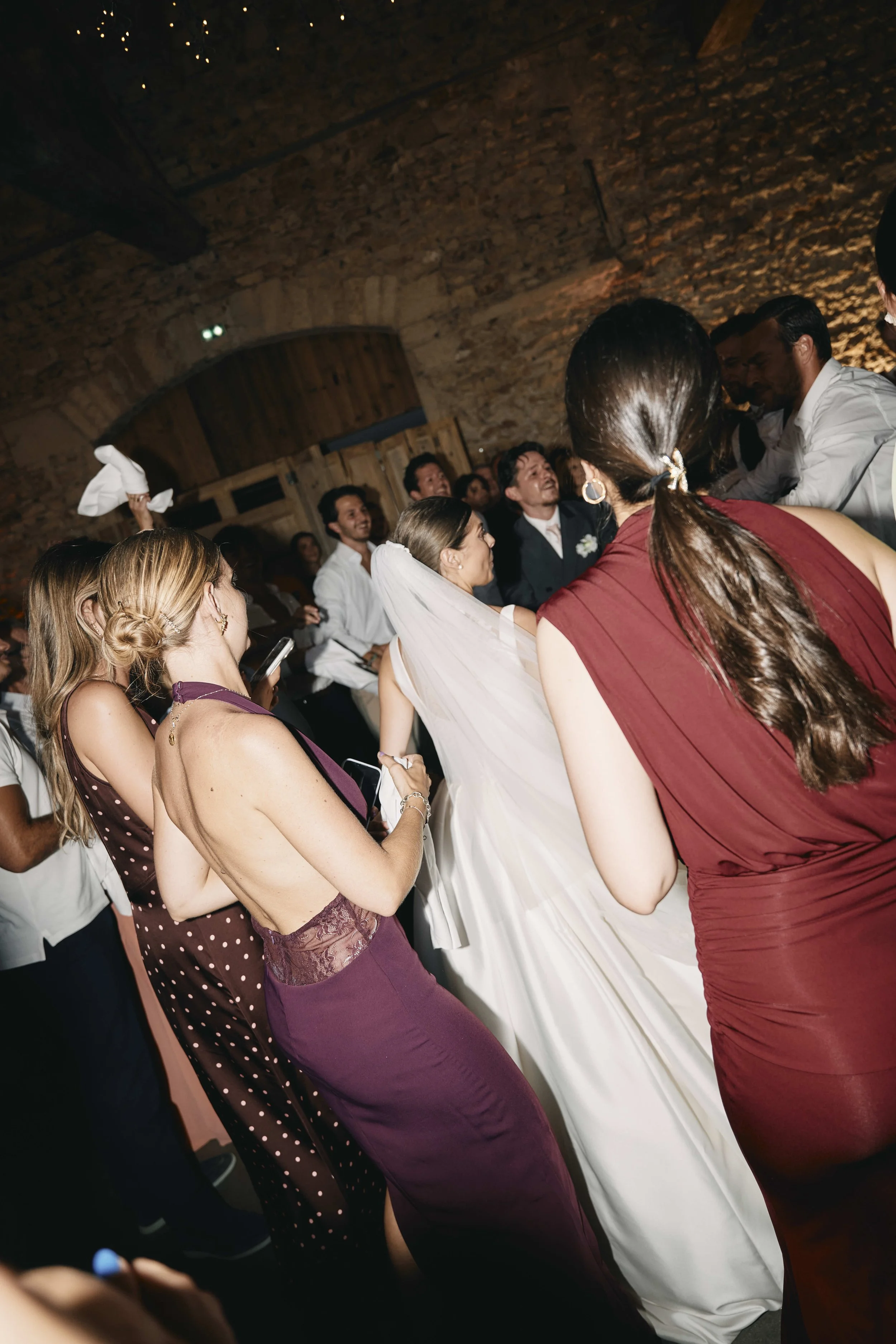 A wedding celebration with guests gathered around, some taking photos and others dancing in a rustic venue with brick walls and wooden accents.