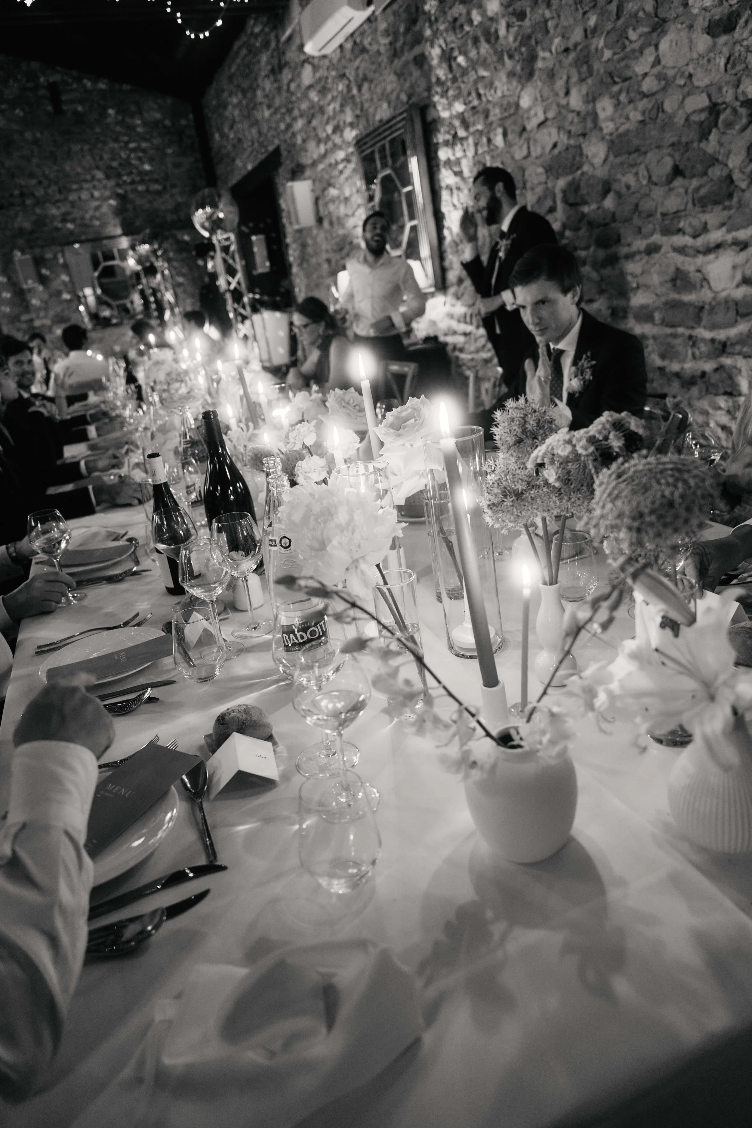A black-and-white photo of a formal dinner party with a long table set with flowers, candles, wine bottles, glasses, and place settings. Several guests are seated, and some are standing and talking. The background features a rustic stone wall and lar
