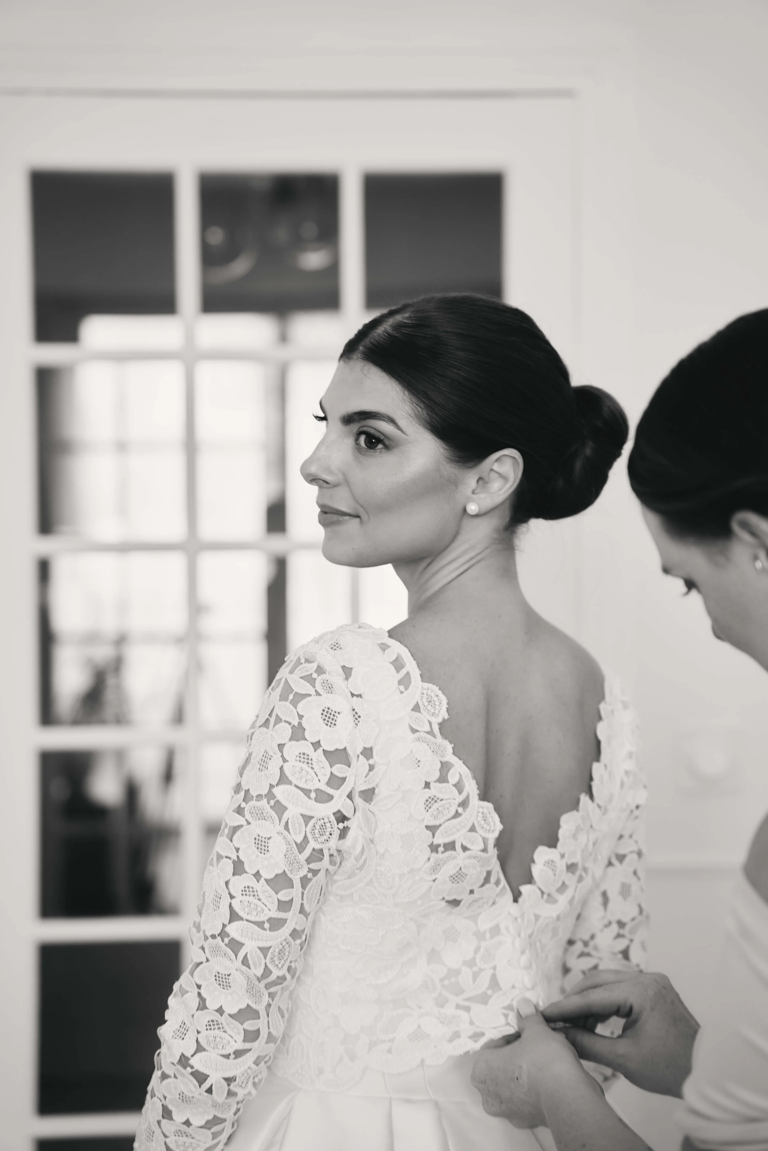 Black and white photo of a woman in a lace wedding dress, looking to the side, having her dress fitted by a person whose hands are visible.