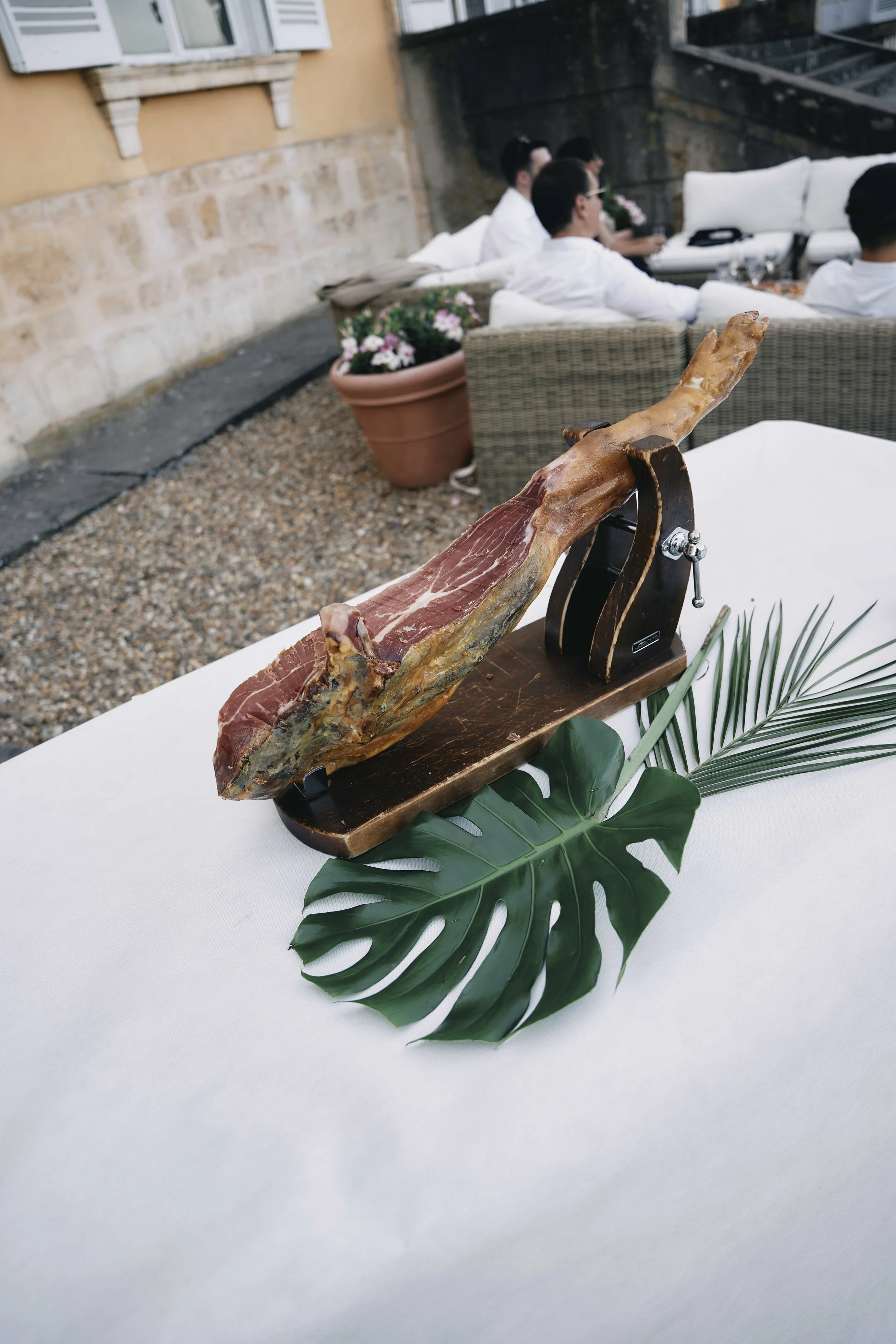 A leg of prosciutto on a wooden stand with a tropical leaf underneath, set on a white table.