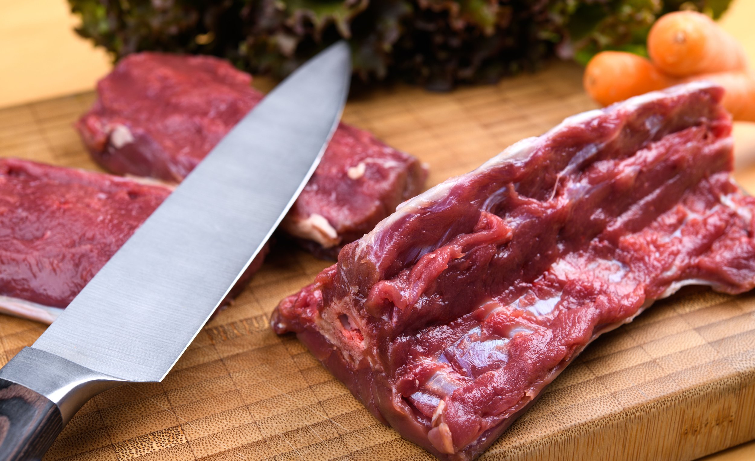 Raw beef on a wooden cutting board with a large knife and some vegetables in the background.
