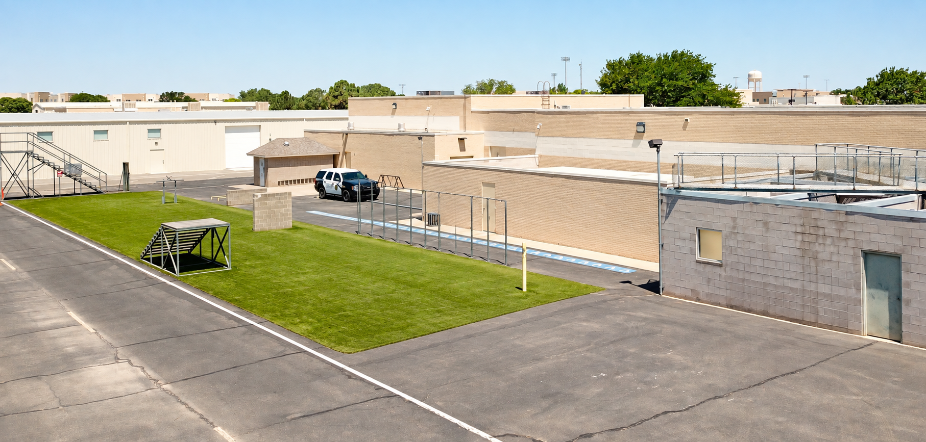 View of a school playground with a grassy area, a small set of stairs, and a basketball hoop, surrounded by school buildings, parking lot with a black SUV, and blue sky.