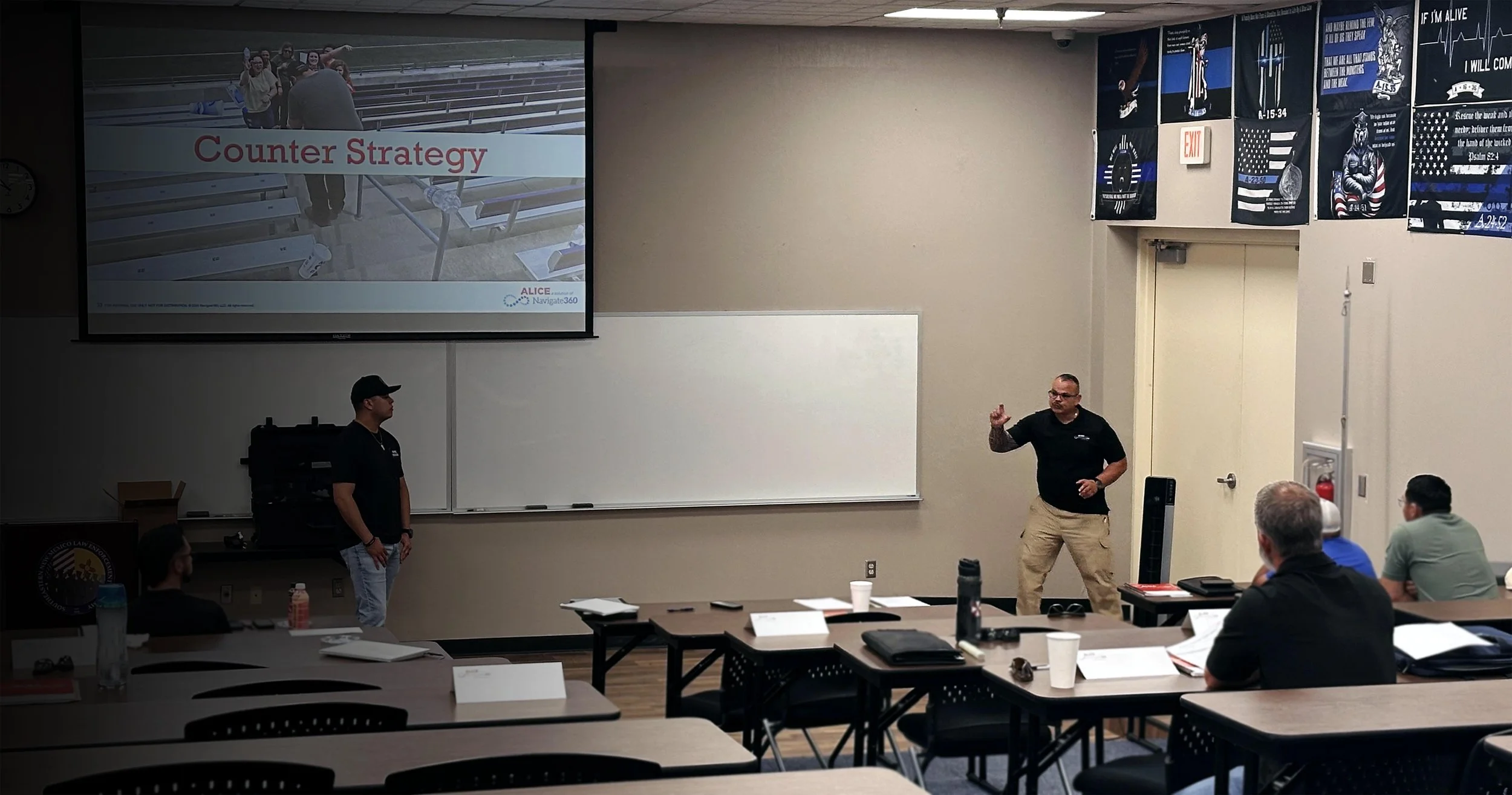 A classroom or conference room with a presentation on a large screen titled 'Counter Strategy.' A man is standing next to the screen, pointing, while another standing person is nearby. Several people are seated at tables, some taking notes, listening to the presentation.