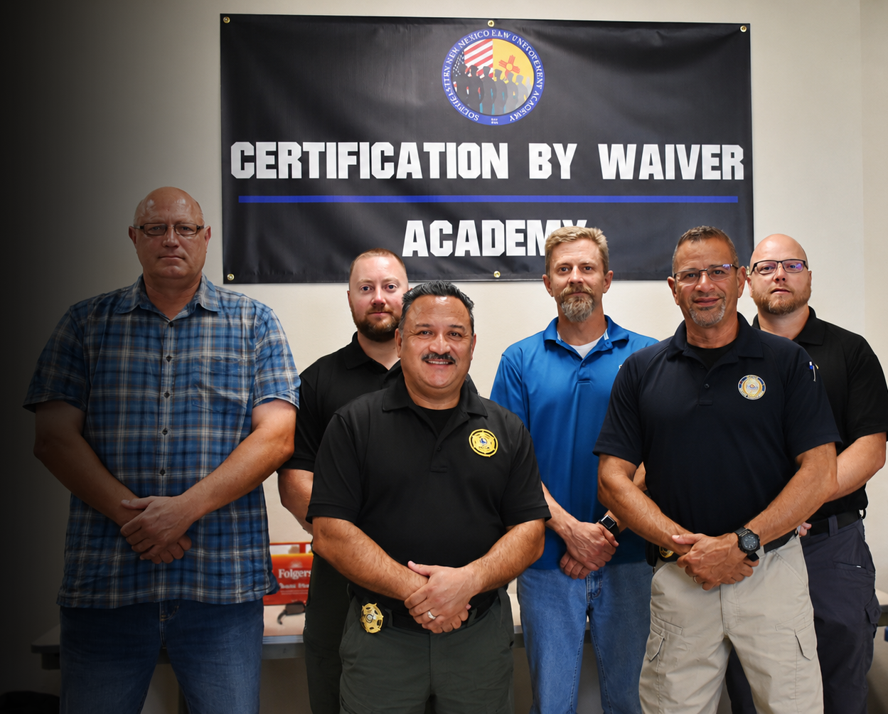 Group of six men standing in front of a black banner that reads 'CERTIFICATION BY WAIVER ACADEMY' with an emblem at the top. The men are wearing casual clothing, some with badges or patches, and are posing for the photo indoors.