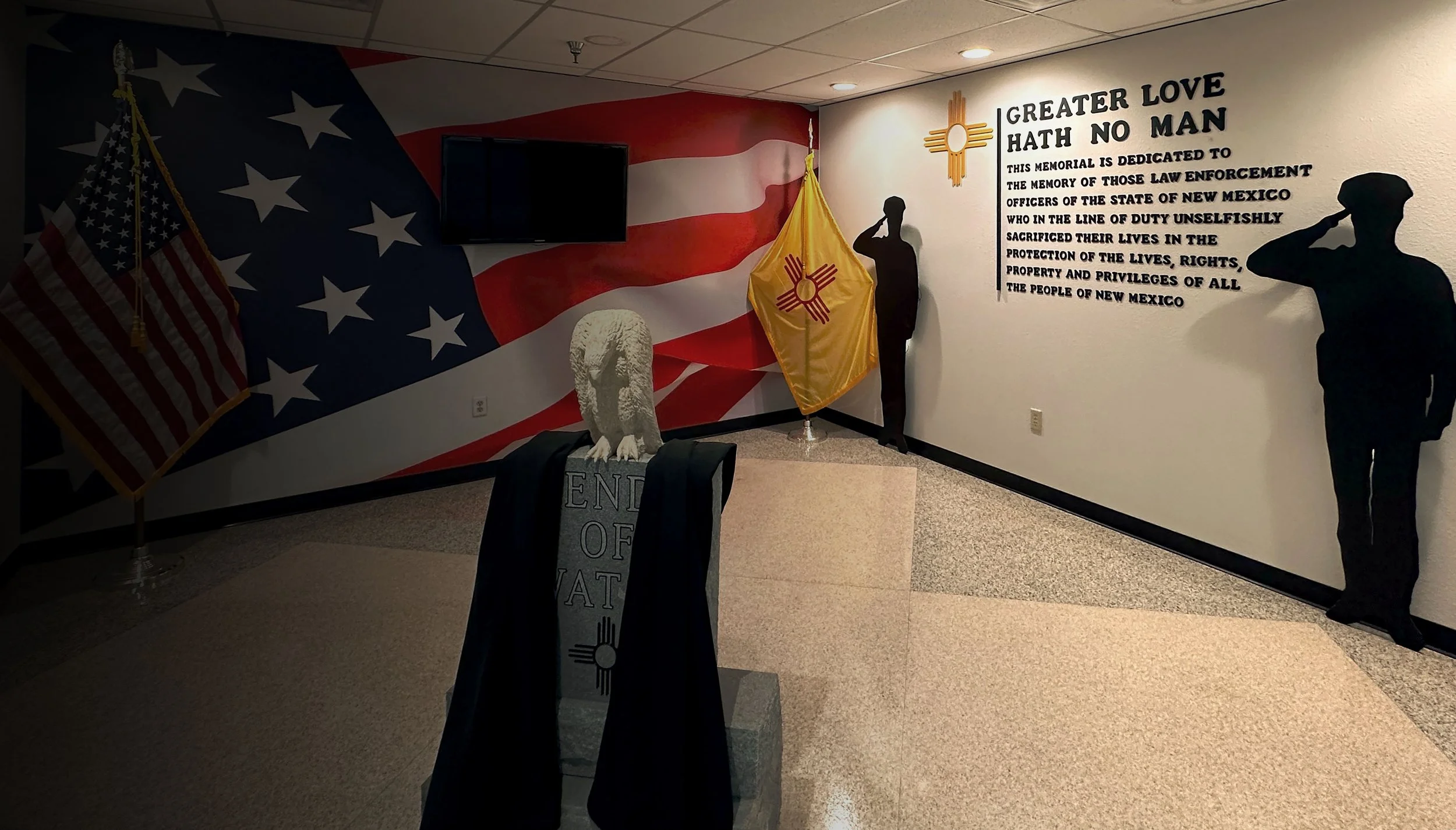 Memorial display featuring the American flag, a yellow and red flag, a stone eagle statue, and silhouettes of saluting soldiers on the wall. The wall has a quote honoring law enforcement officers from New Mexico.