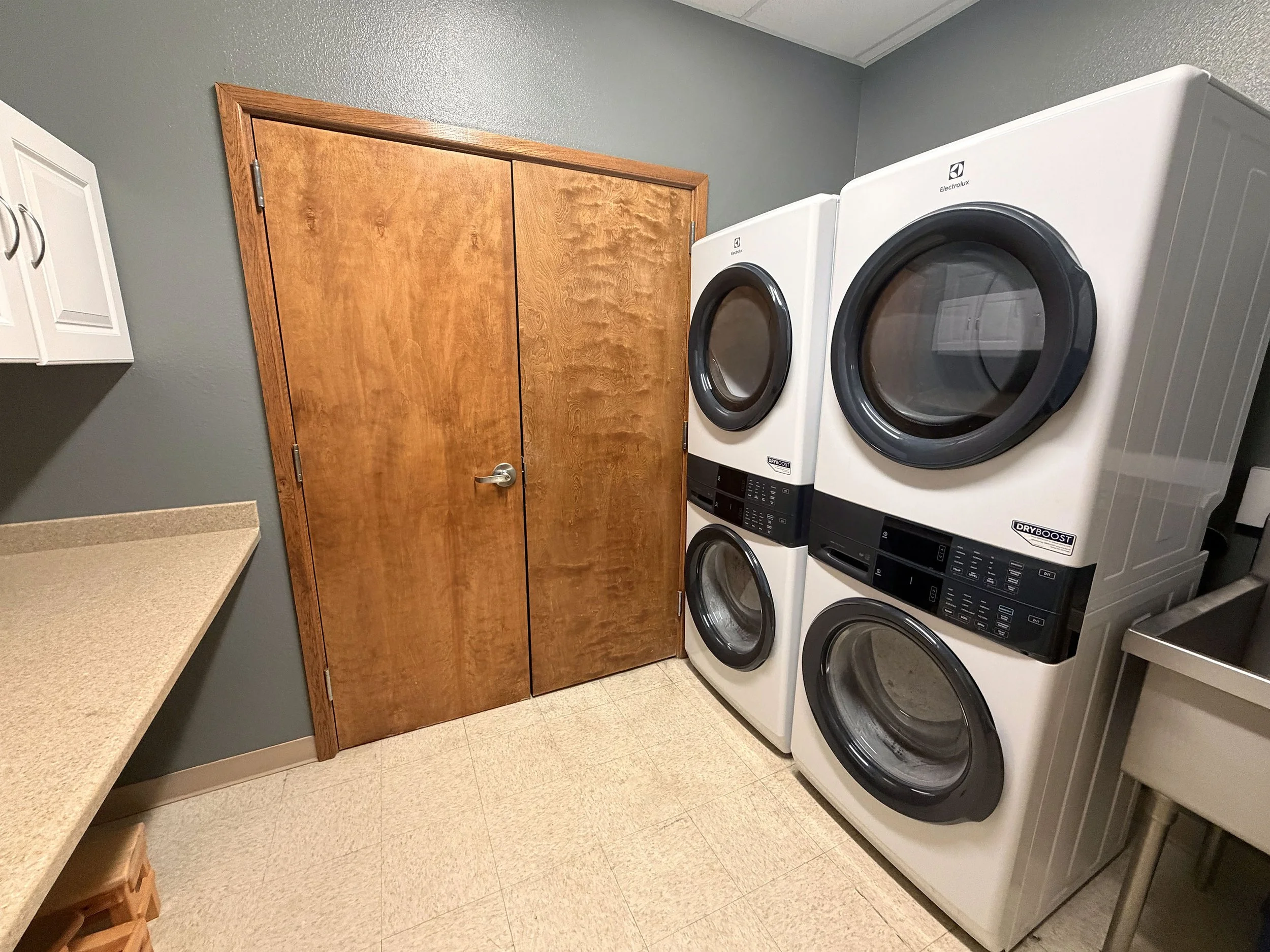 A laundry room with a wooden door, gray walls, beige countertop, and white front-loading washer and dryer stacked on the right.