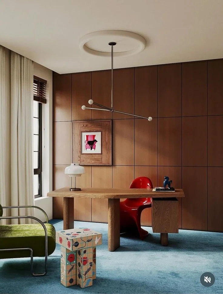 A modern home office with wooden paneled wall, a desk, a red chair, a green chair, a colorful patterned small table, and a window with curtains.