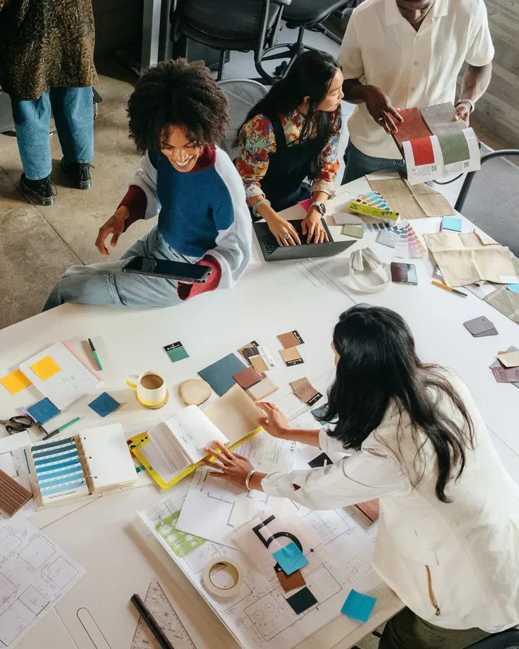 Group of diverse women working on a design project at a table with paint chips, color samples, notebooks, and a laptop, in a casual office setting.