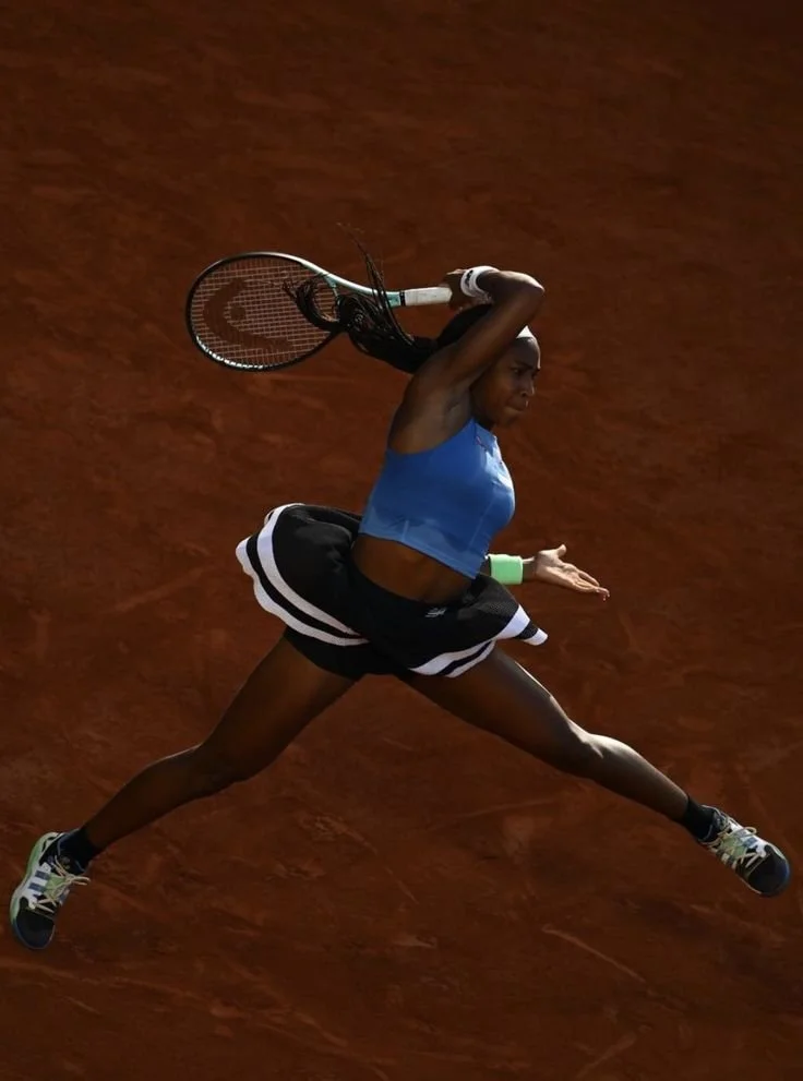 A female tennis player in a blue sports bra and black athletic shorts is jumping on a clay tennis court, holding a tennis racquet above her head.