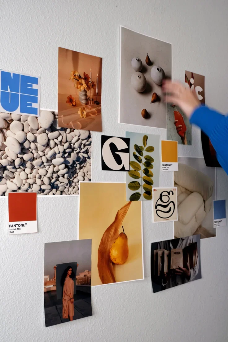 A wall decorated with various photos, postcards, and color samples, including images of stones, flowers, pears, a woman, and abstract art, with a person's hand reaching toward one of the photos.