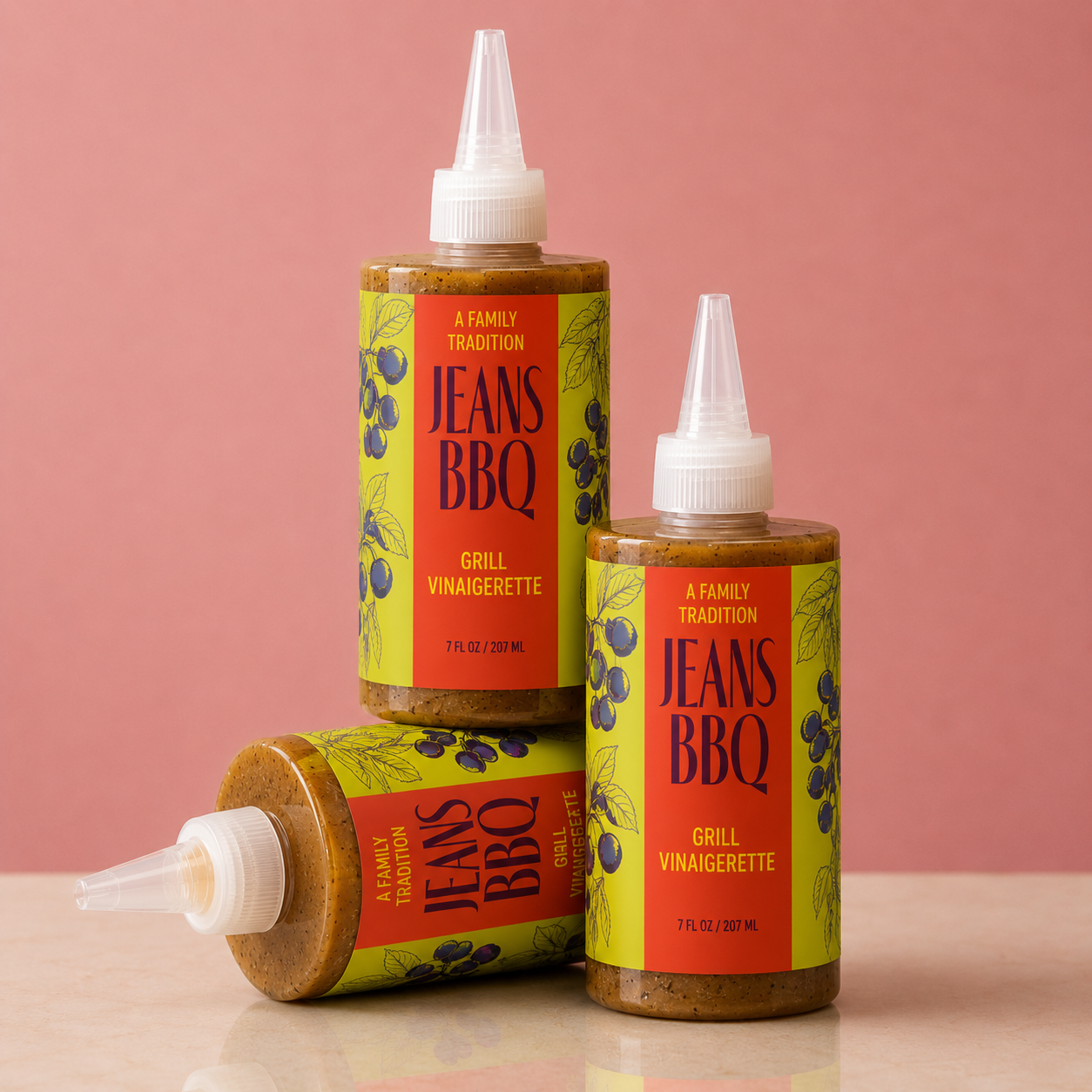 Three bottles of Jeans BBQ Grill Vinaigrette sauce with yellow and red labels, blueberries, and leaves on a pink background, one lying down and two standing.
