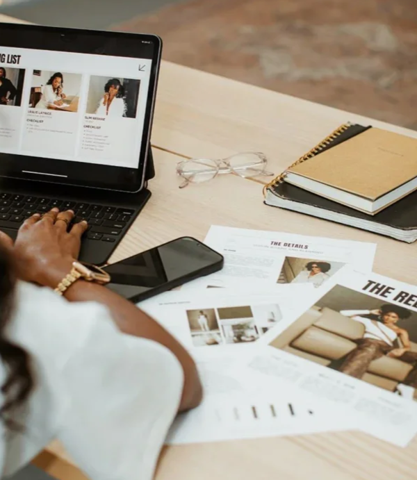 Person working at a desk with a tablet, smartphone, glasses, notebooks, and printed documents about fashion and models.