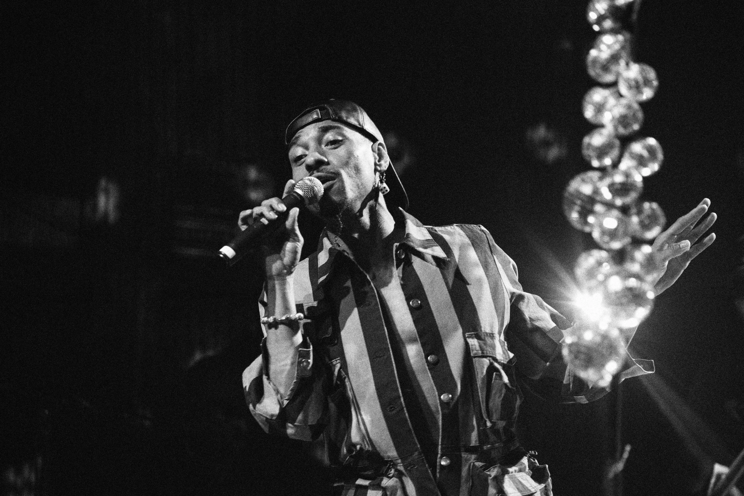 Durrand Brenarr singing into a microphone with a beaded bracelet on his wrist, wearing a plaid shirt and a hat backward, in a dimly lit setting.